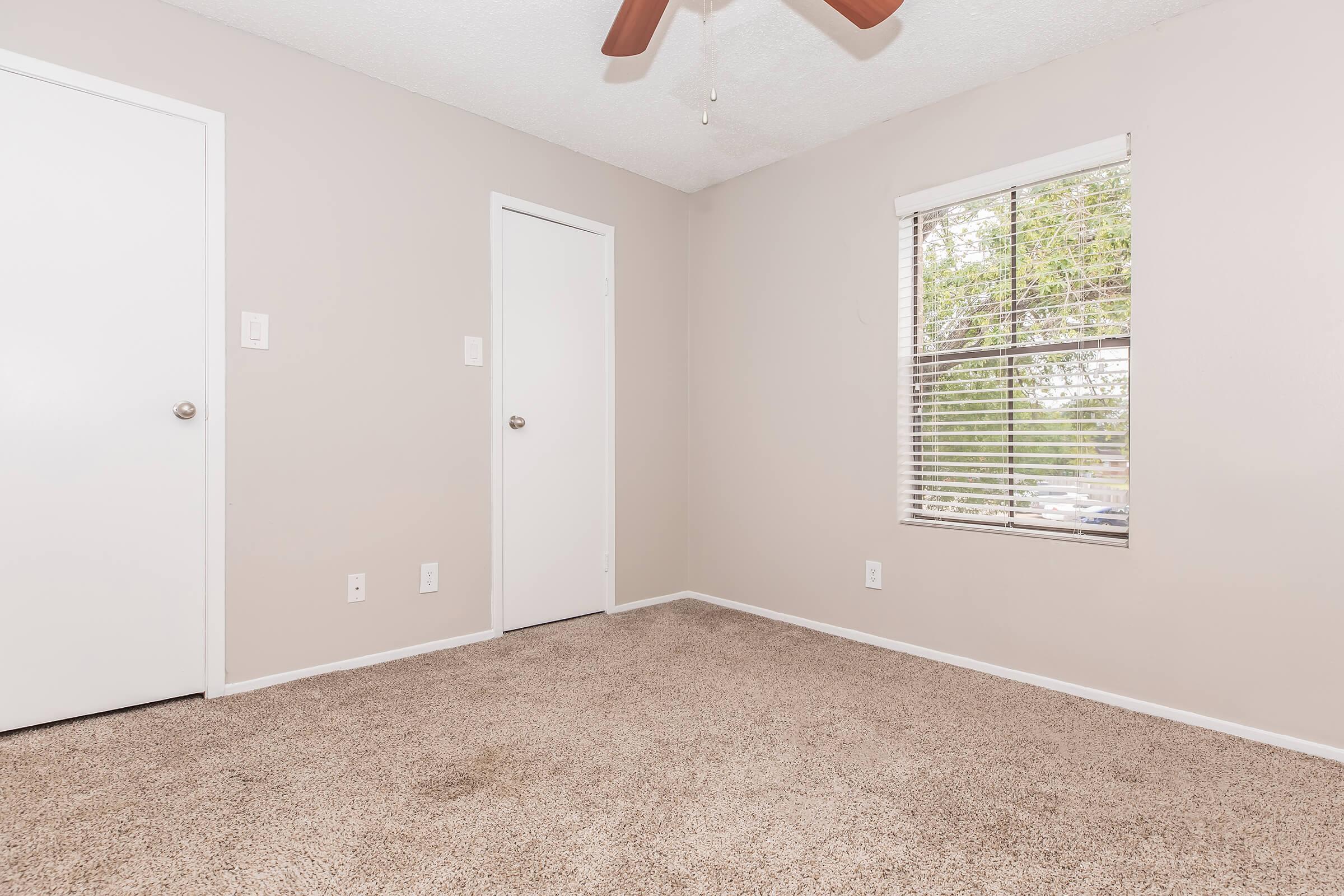 Empty room with light beige walls and carpet. A ceiling fan is visible. There are two doors: one leading to a closet and the other is a standard door. A window with blinds allows natural light. The space has a clean and simple design, suitable for various uses.