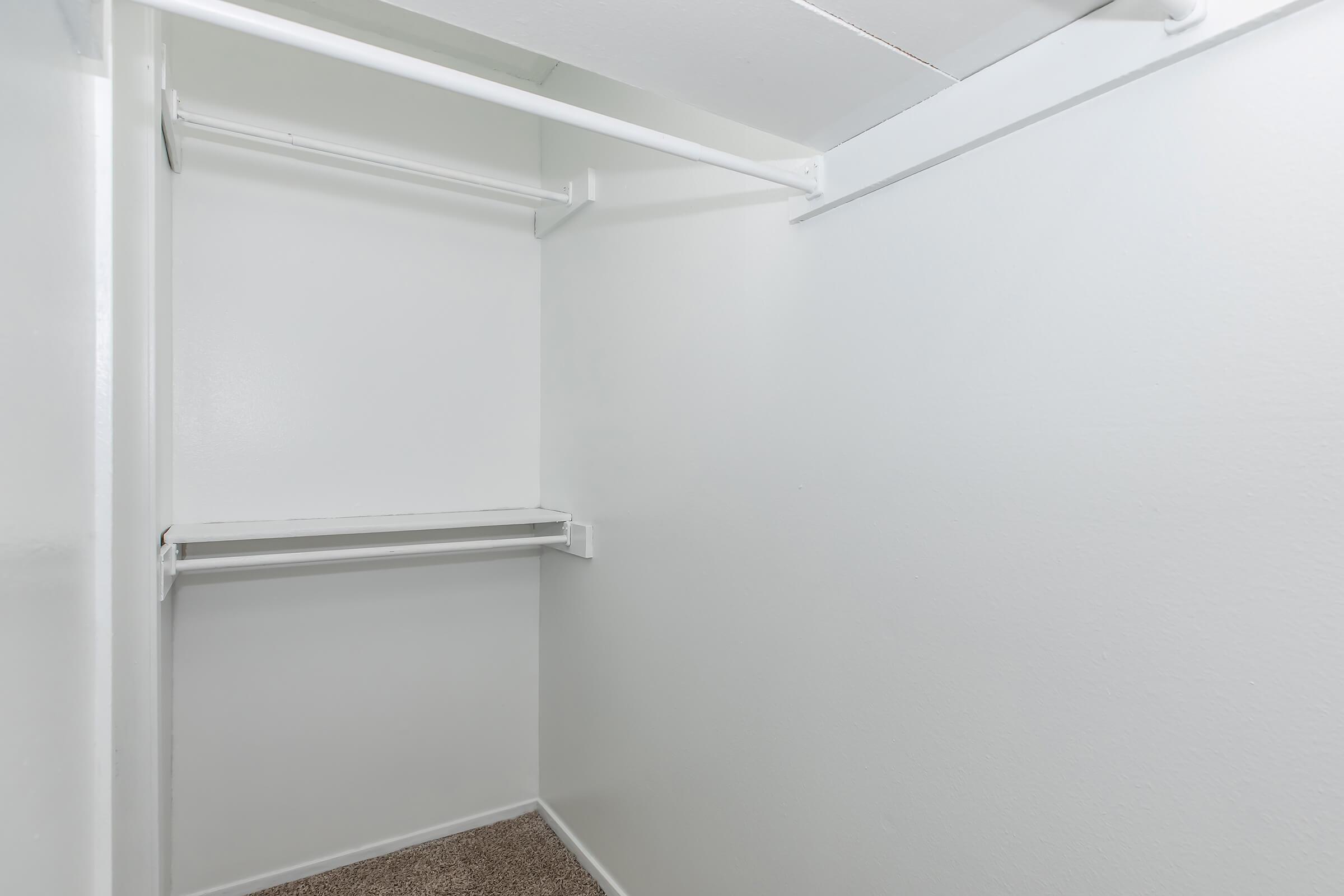 An empty closet with white walls and two hanging rods. The floor is carpeted. There are no shelves or stored items visible, creating a spacious and unobstructed interior.