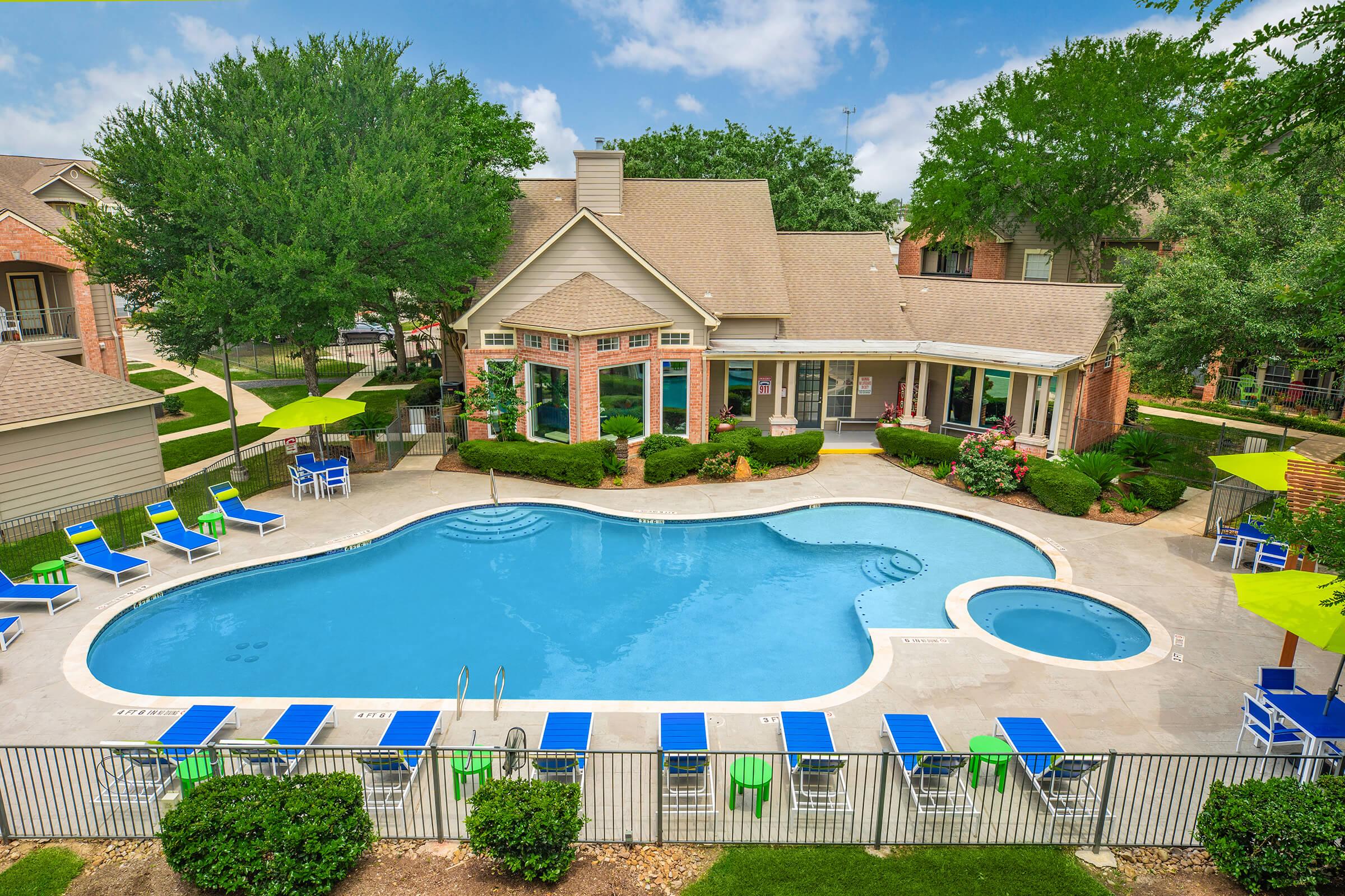 Aerial view of a residential pool area featuring a large, curvy swimming pool surrounded by lounge chairs and green umbrellas. The pool is bordered by well-maintained gardens and a modern building with large windows. Lush trees and shrubs enhance the inviting atmosphere of the outdoor space.