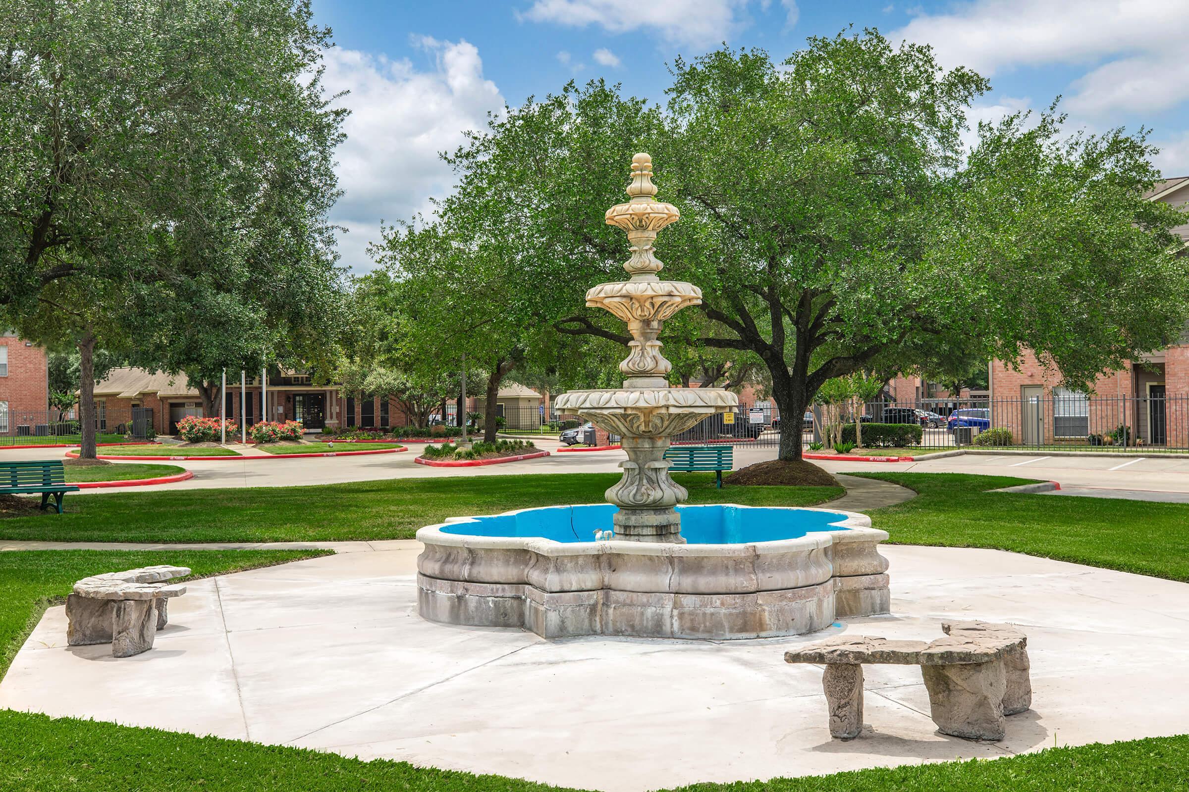A decorative stone fountain sits at the center of a grassy area, surrounded by trees. The fountain features multiple tiers and is filled with blue water. Two stone benches are placed nearby on a concrete surface, with a peaceful park setting in the background. Bright blue sky and fluffy clouds add to the serene atmosphere.