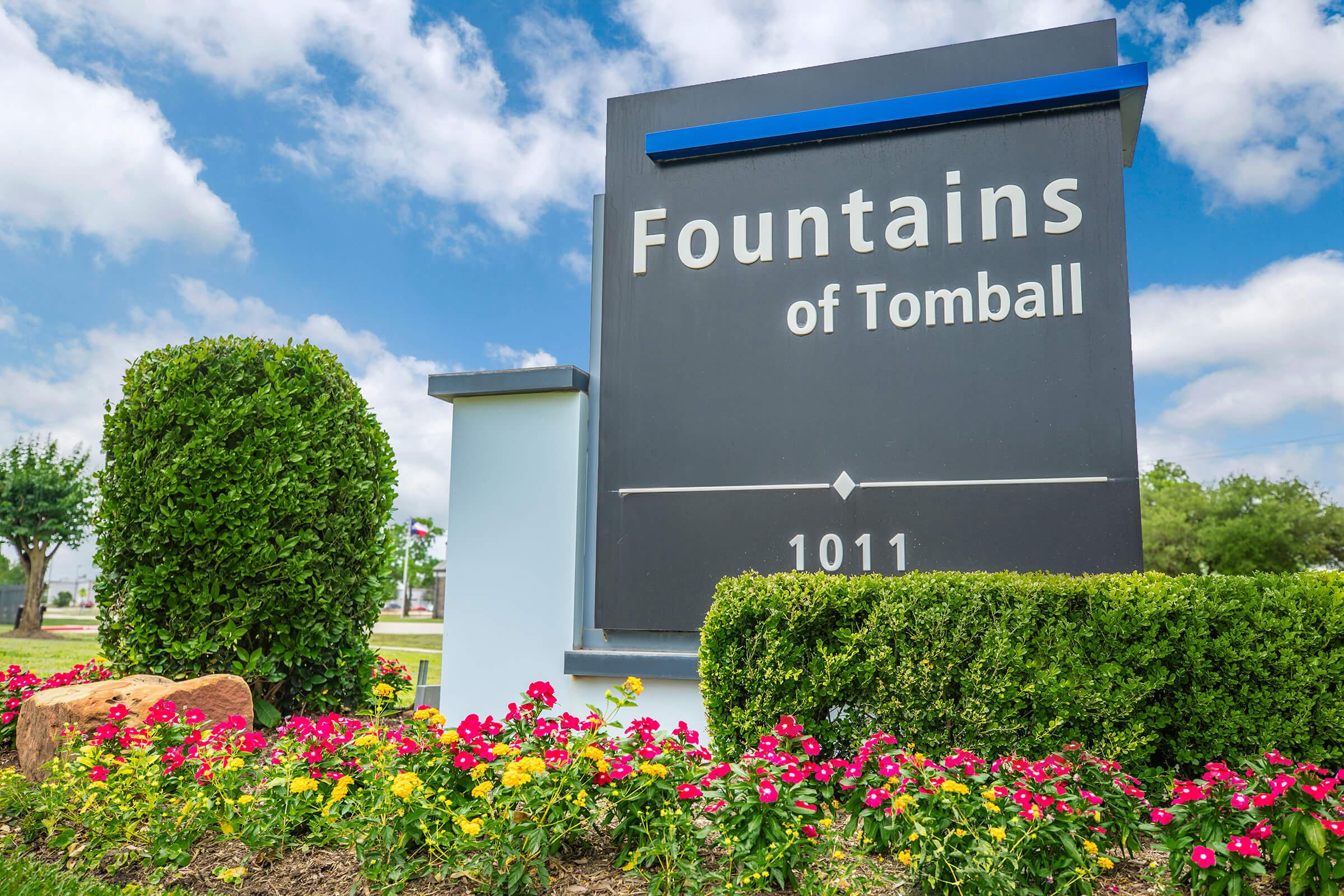 Sign for "Fountains of Tomball" with the address 1011, surrounded by colorful flowers and manicured bushes under a partly cloudy sky.