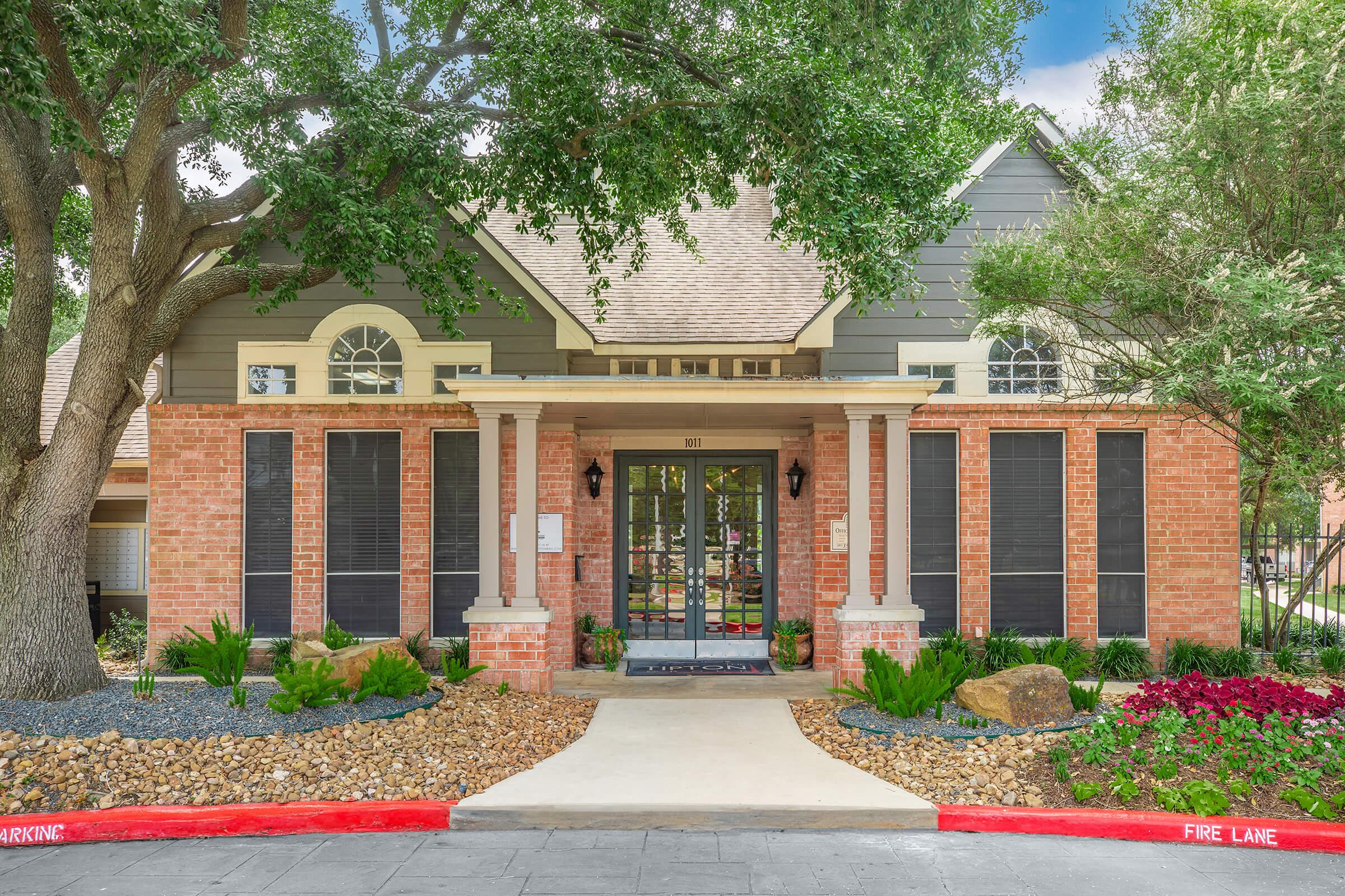 A charming brick and gray house with a covered porch, surrounded by lush greenery and landscaped stones. The entrance features large windows and decorative shutters, with a clear path leading to the door. The property includes well-maintained shrubs and a red curb marking a fire lane.