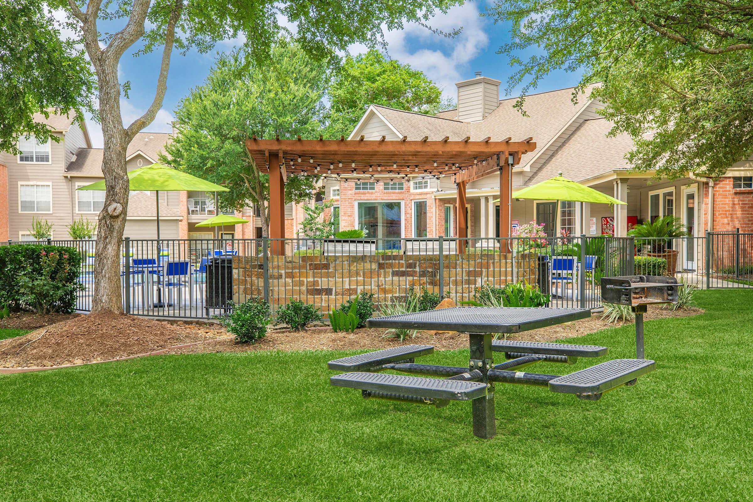 A landscaped outdoor area featuring a picnic table on green grass, surrounded by various plants. There is a stone patio with a pergola in the background, along with bright green umbrellas over blue chairs. The setting is part of an apartment complex, with residential buildings visible nearby.