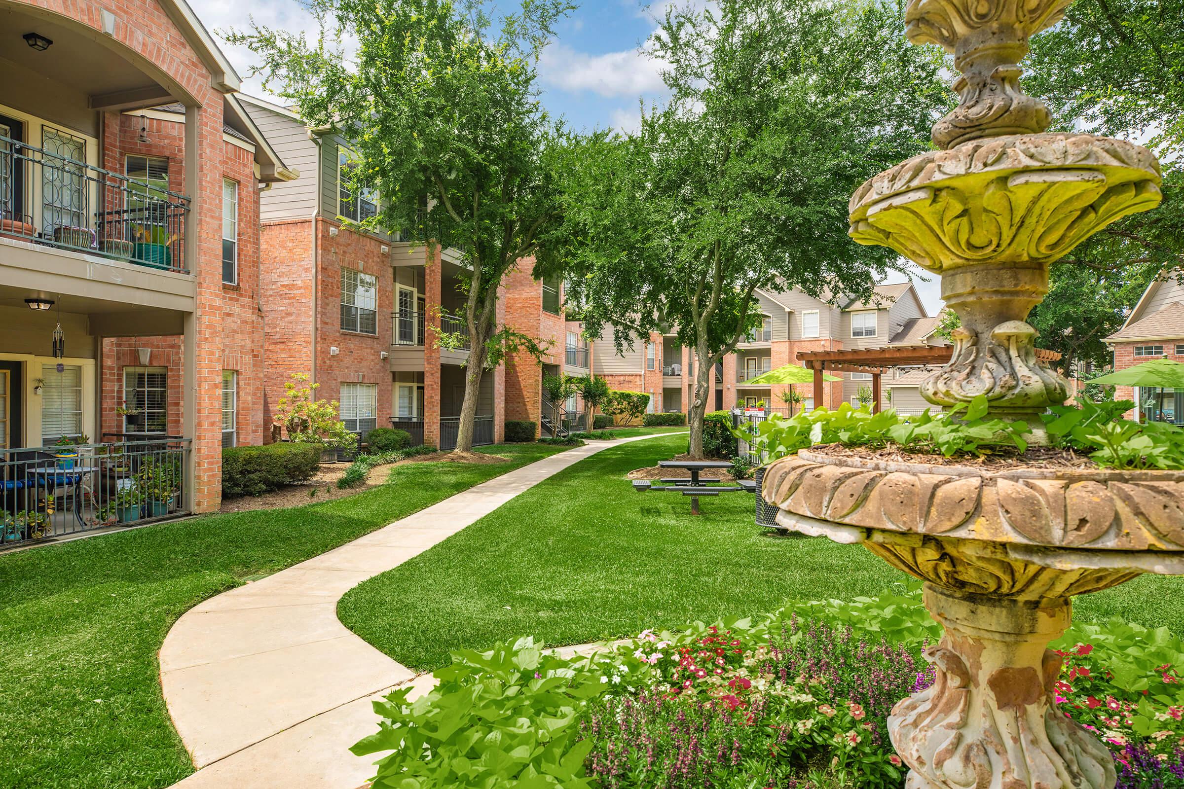 A landscaped courtyard at an apartment complex, featuring a stone fountain surrounded by greenery. The pathway winds through lush grass and flower beds, with brick buildings and trees visible in the background under a blue sky.