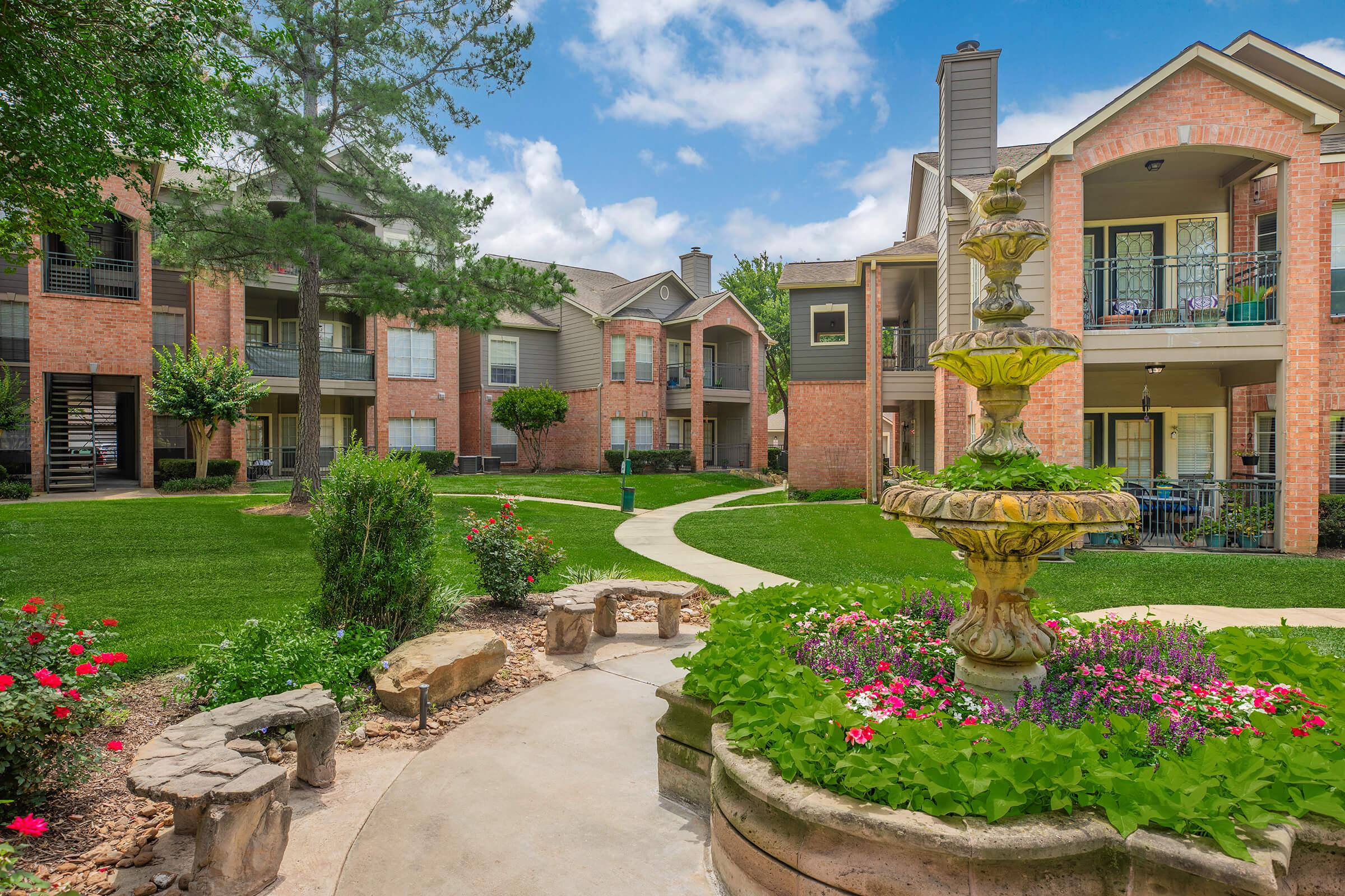 A serene courtyard featuring a decorative fountain surrounded by colorful flowers and neatly landscaped greenery. In the background, brick apartment buildings are visible under a partly cloudy sky, creating a peaceful residential atmosphere. Pathways wind through the garden, inviting residents to stroll.