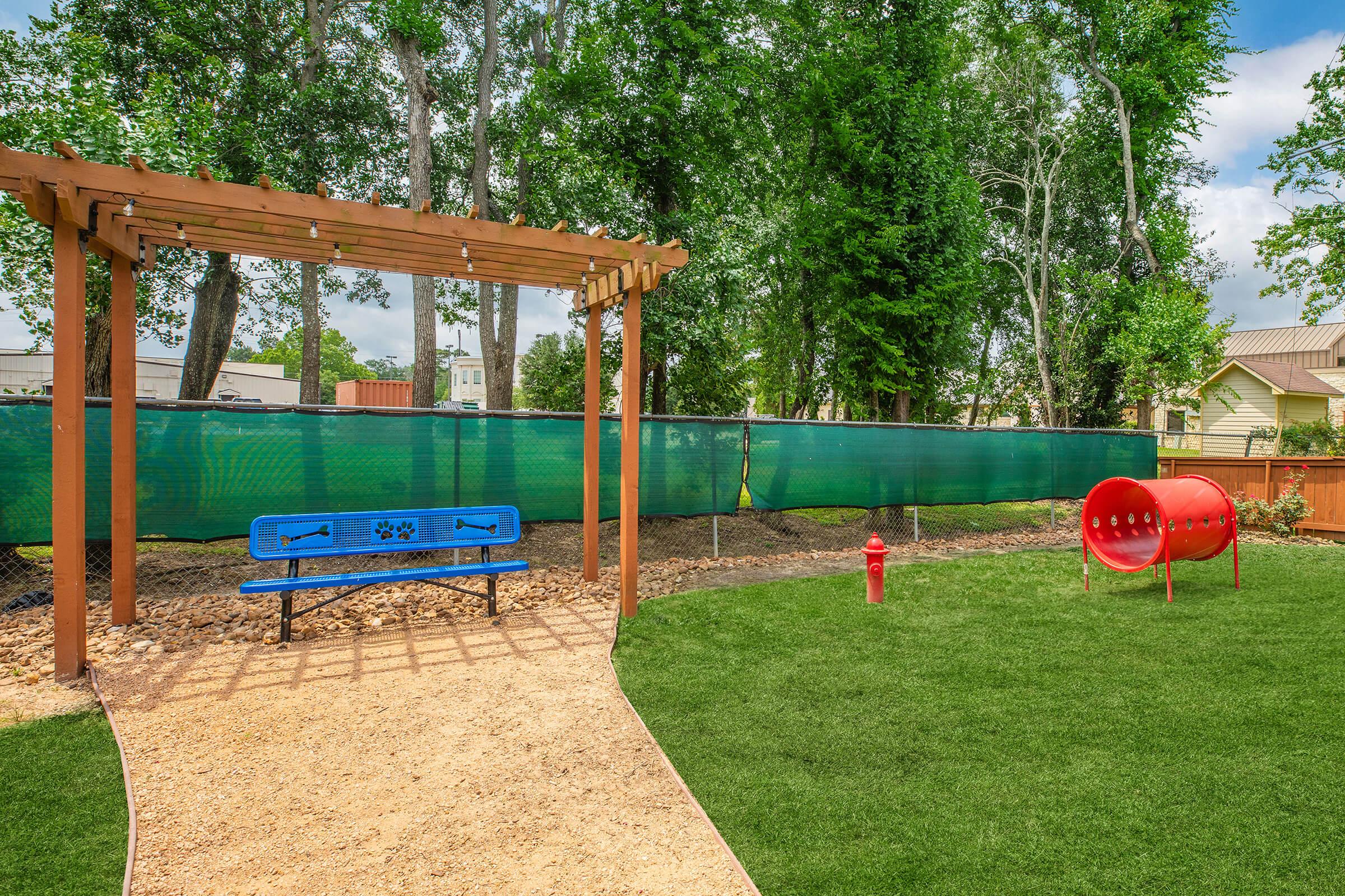 A pet-friendly park area featuring a blue bench under a wooden pergola, a red dog tunnel, and a fire hydrant on a grassy lawn. The space is enclosed by a green fence and surrounded by trees, creating a welcoming outdoor environment for dogs and their owners.
