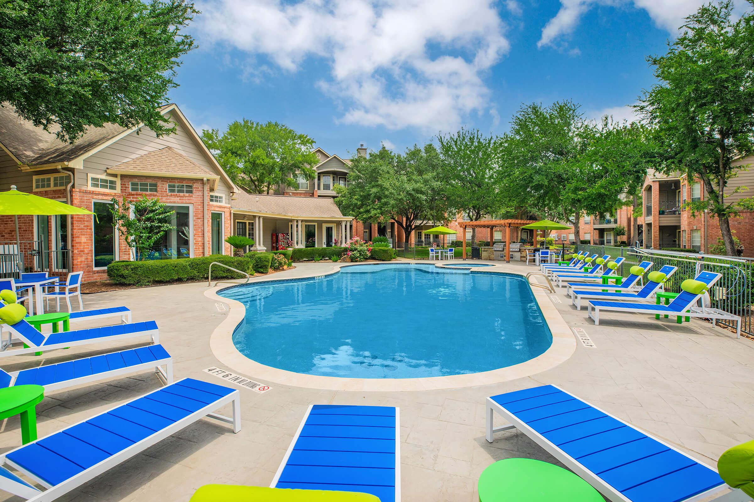 A clear blue swimming pool surrounded by lounge chairs in bright blue and green. Lush greenery and trees provide shade around the pool area, with a few umbrellas nearby. Residential buildings are visible in the background under a partly cloudy sky.