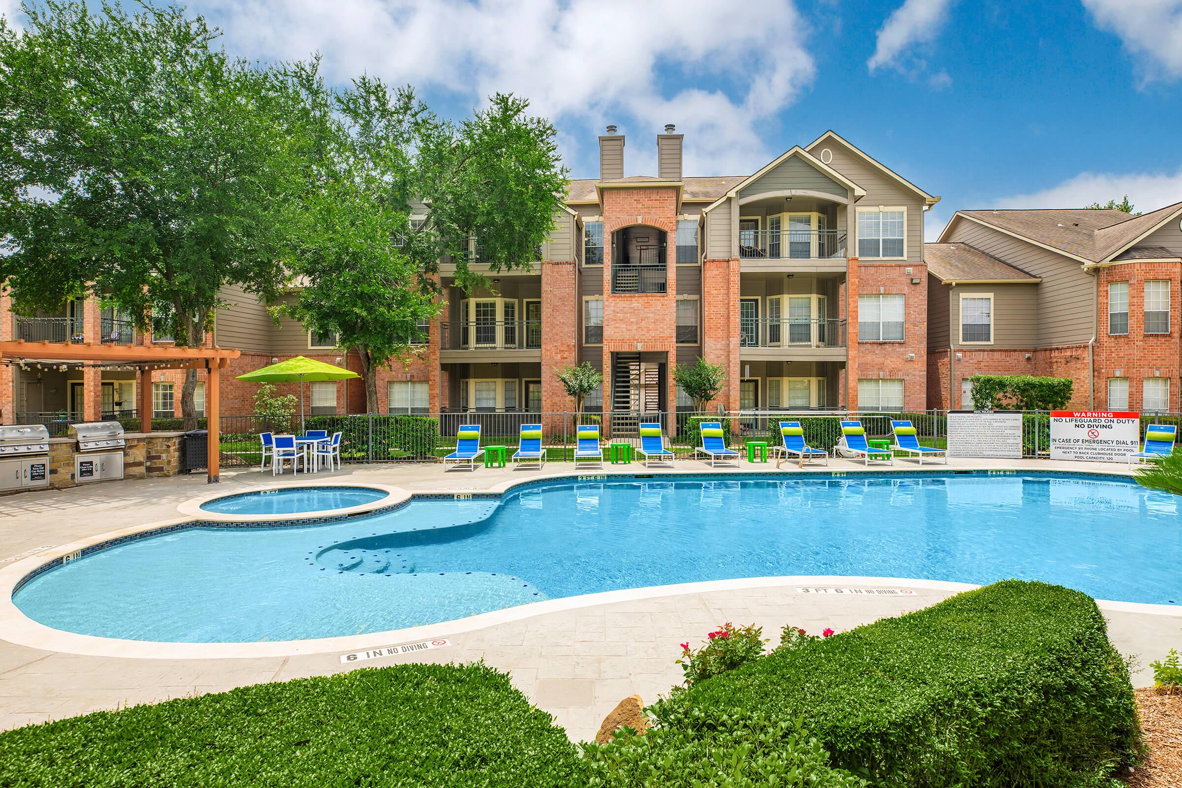 A swimming pool area surrounded by greenery, featuring lounge chairs and umbrellas. In the background, there are residential buildings with balconies and large windows. The scene is bright and sunny, with a clear blue sky. Outdoor barbecue grills are also visible near the pool.
