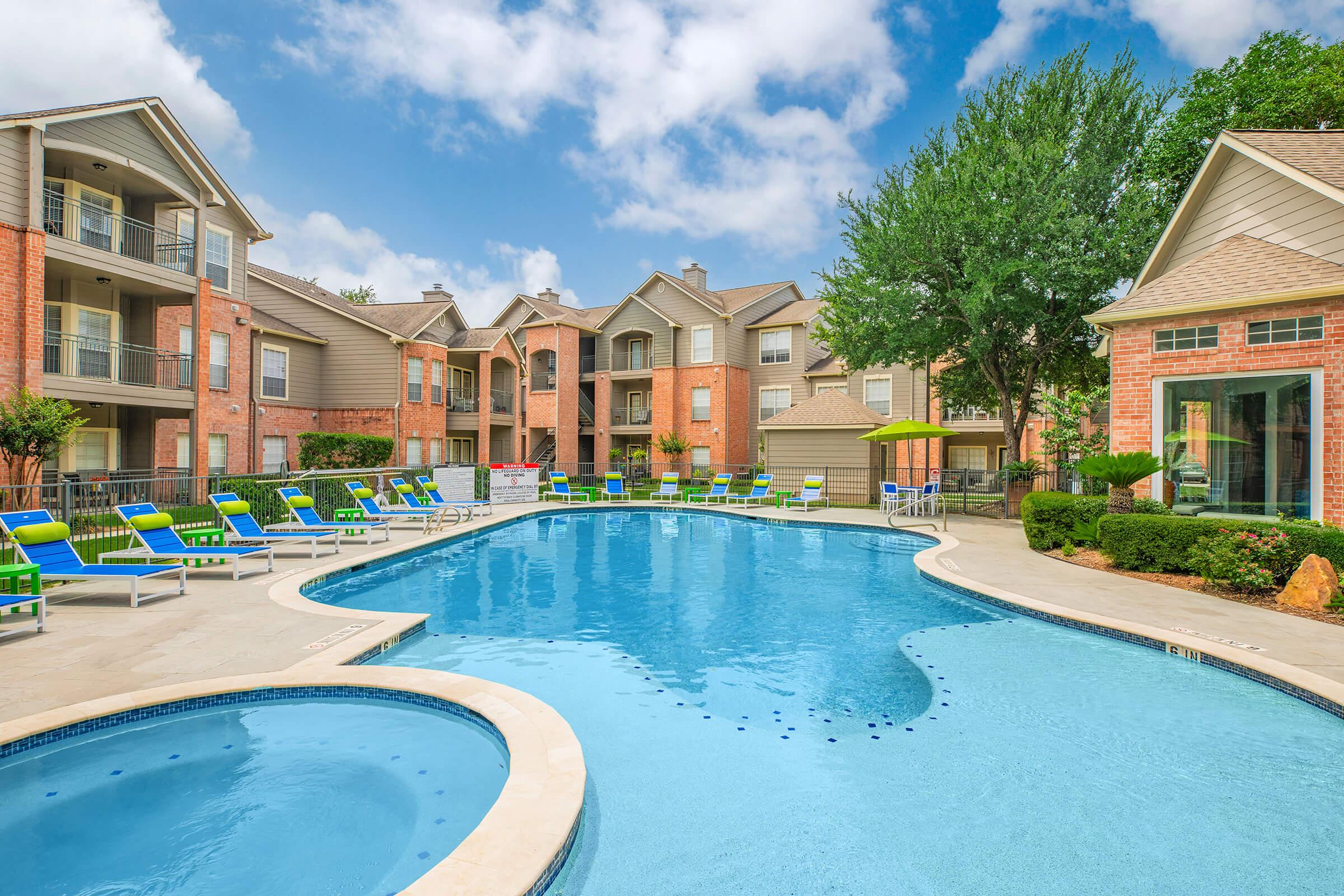 A large swimming pool surrounded by lounge chairs and umbrellas, situated in a residential area. Nearby, there are brick apartment buildings with balconies and well-maintained landscaping. The sky is partly cloudy, creating a bright, inviting atmosphere.
