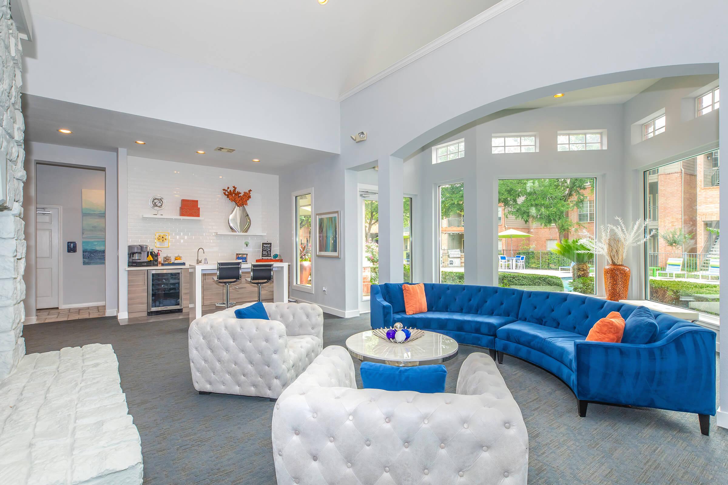 A bright and modern common area featuring a blue velvet sofa and two tufted white chairs surrounding a round table. The space includes large windows with a view of greenery outside, a kitchenette with sleek appliances, and decorative elements like artwork and plants.