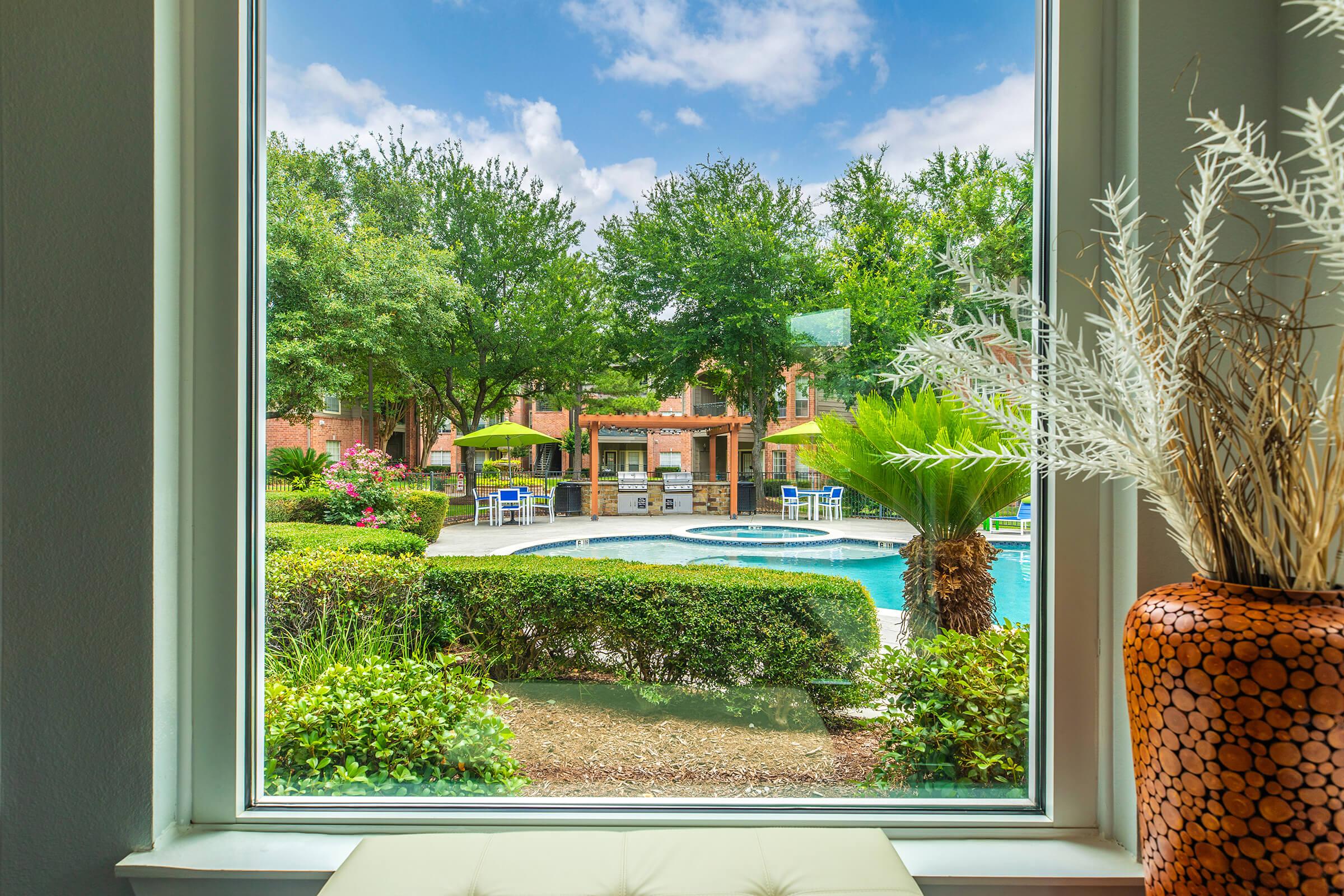 View from a window overlooking a pool area surrounded by lush greenery and umbrellas. The scene features palm trees, flower beds, and a cozy outdoor seating area, creating a relaxing atmosphere under a partly cloudy sky.