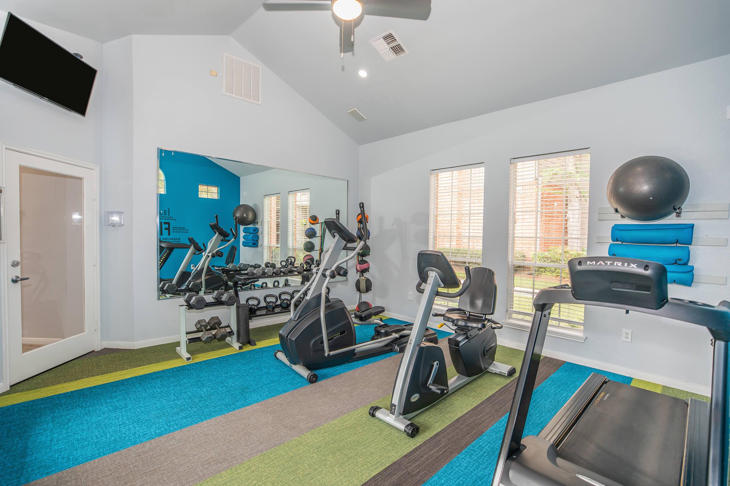 A bright and modern gym space featuring exercise equipment such as treadmills and stationary bikes, free weights, and a large mirror. The room is decorated with colorful carpeting and has natural light coming through several windows, creating an inviting atmosphere for workouts.