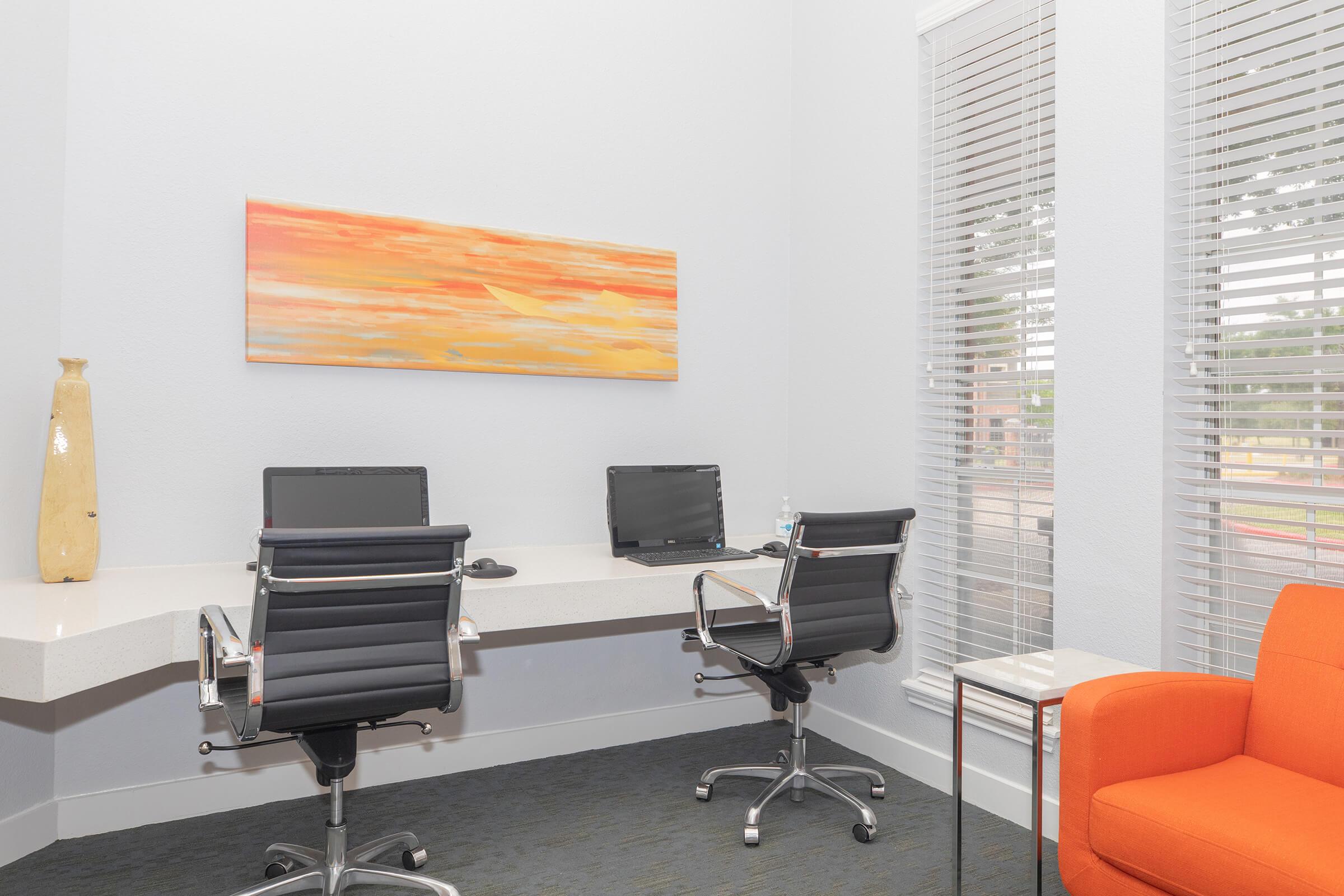 A modern office space featuring two sleek black chairs at a white desk with two computers. There is an orange armchair in the corner and a colorful abstract painting on the wall. The room has large windows with blinds letting in natural light, creating a bright and inviting atmosphere.