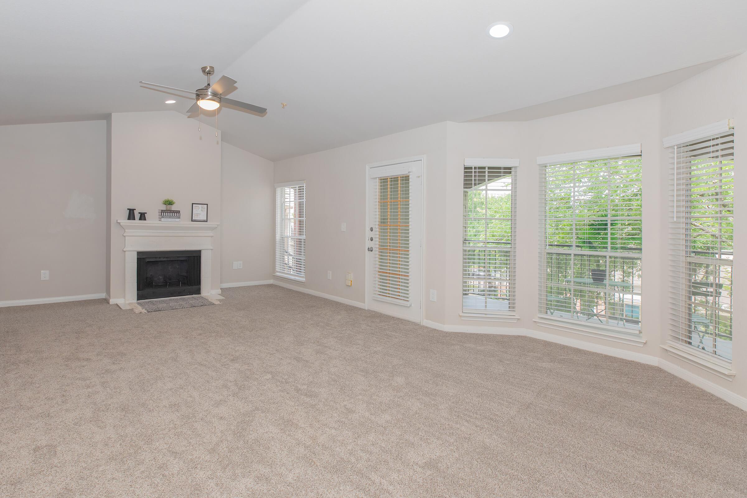 Spacious living room featuring light-colored walls, a ceiling fan, and a cozy fireplace. Large windows with white blinds allow natural light to fill the room, while plush carpet flooring adds warmth. The area is bright and inviting, with access to outdoor views.