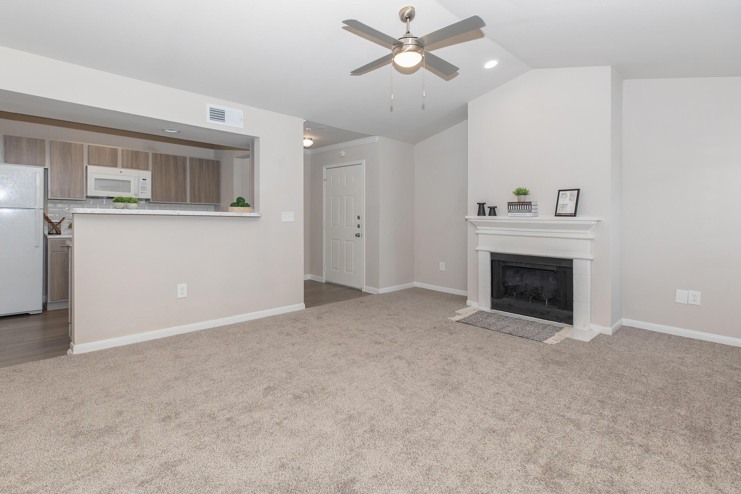 A spacious living area featuring beige walls and carpet. There is a ceiling fan, a white fireplace with a gray mantle, and an open kitchen area with wooden cabinetry. A door leads to the entrance, with a small decorative shelf on the side. Natural light fills the room, creating an inviting atmosphere.