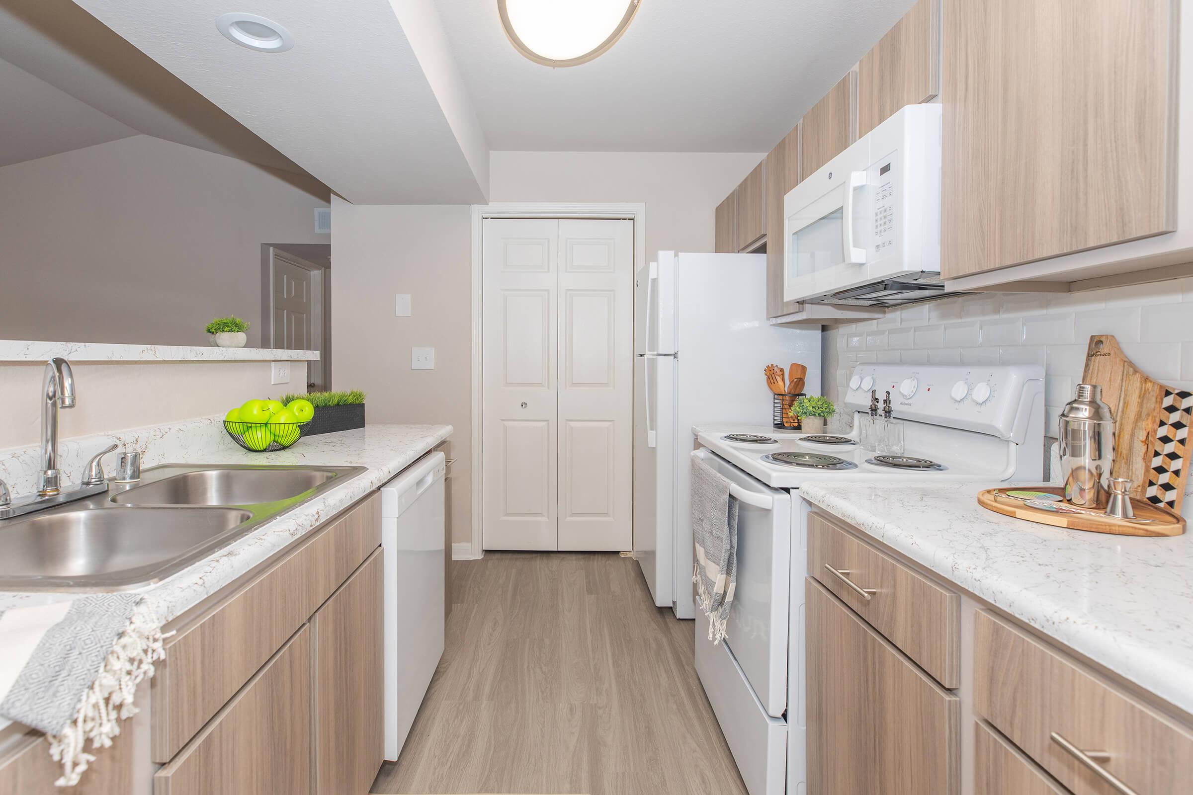 Modern kitchen featuring light wood cabinetry, white appliances, and a white countertop. The space includes a double sink, a microwave, and an oven, with a door leading to a pantry. A bowl of green apples sits on the counter, adding a pop of color to the neutral decor.