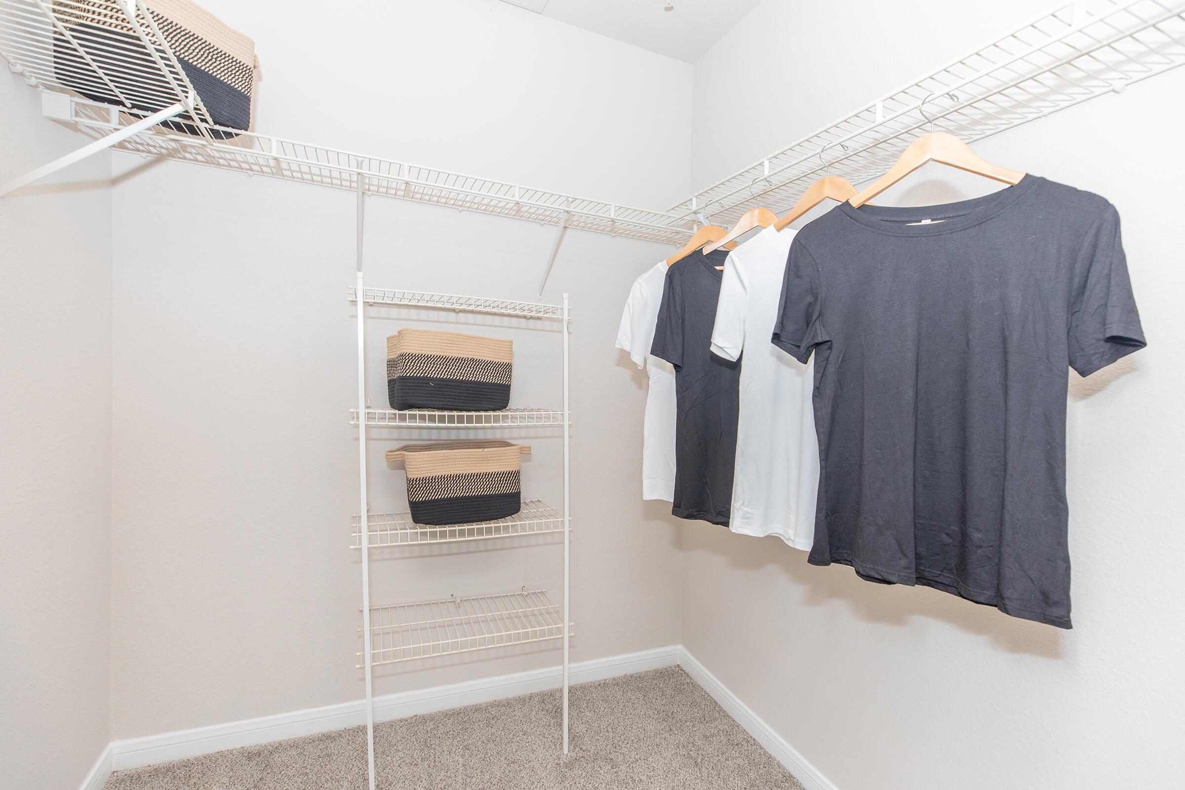 A clean, organized closet featuring a white wire shelving unit. Hanging on the left are several shirts in black and white, while below are stacked woven baskets in a contrasting color. The walls are painted in a light shade, and the floor is carpeted.