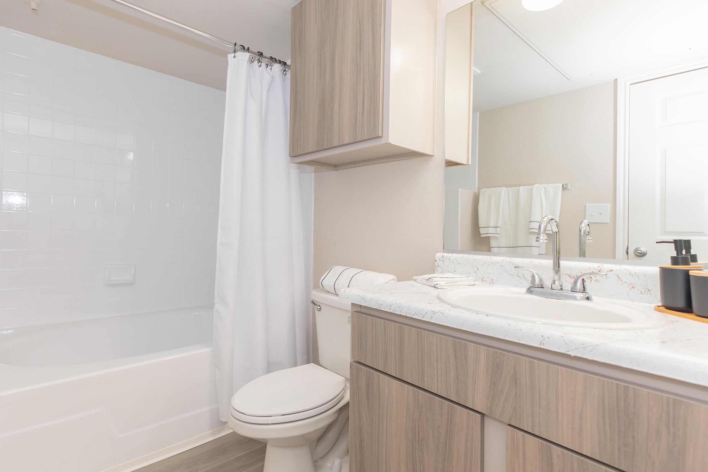 A clean, modern bathroom featuring a white bathtub with an attached shower curtain, a sleek vanity with a sink, and wooden cabinetry. Fresh towels are neatly arranged, and there's a mirror above the sink. The space is well-lit and has a neutral color palette, creating a bright and inviting atmosphere.