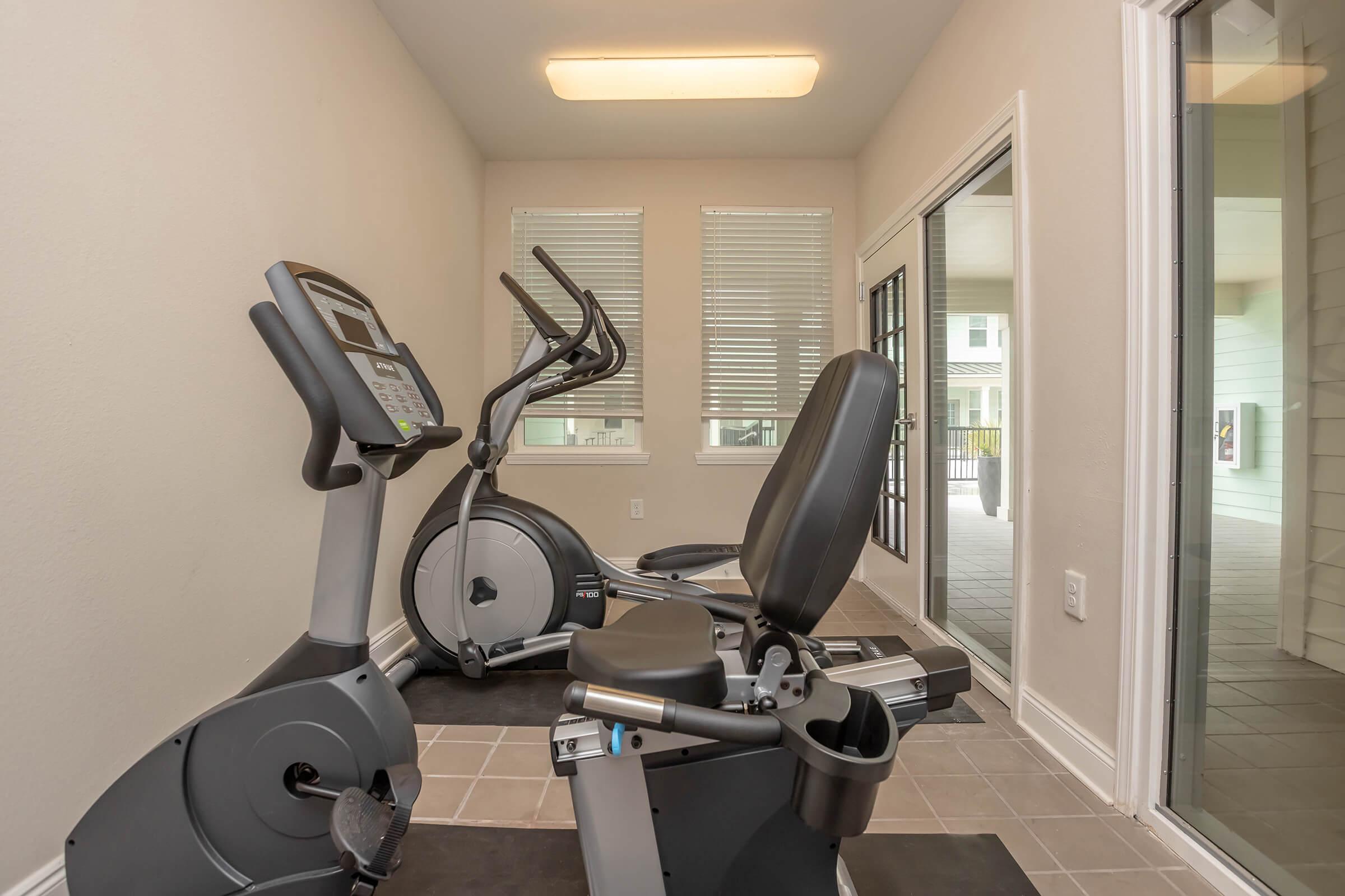 A bright, well-lit fitness room featuring an elliptical machine and a stationary bike, positioned on a tiled floor. Large windows provide views of the outside area, and the minimalist decor emphasizes a clean and modern environment for exercise.