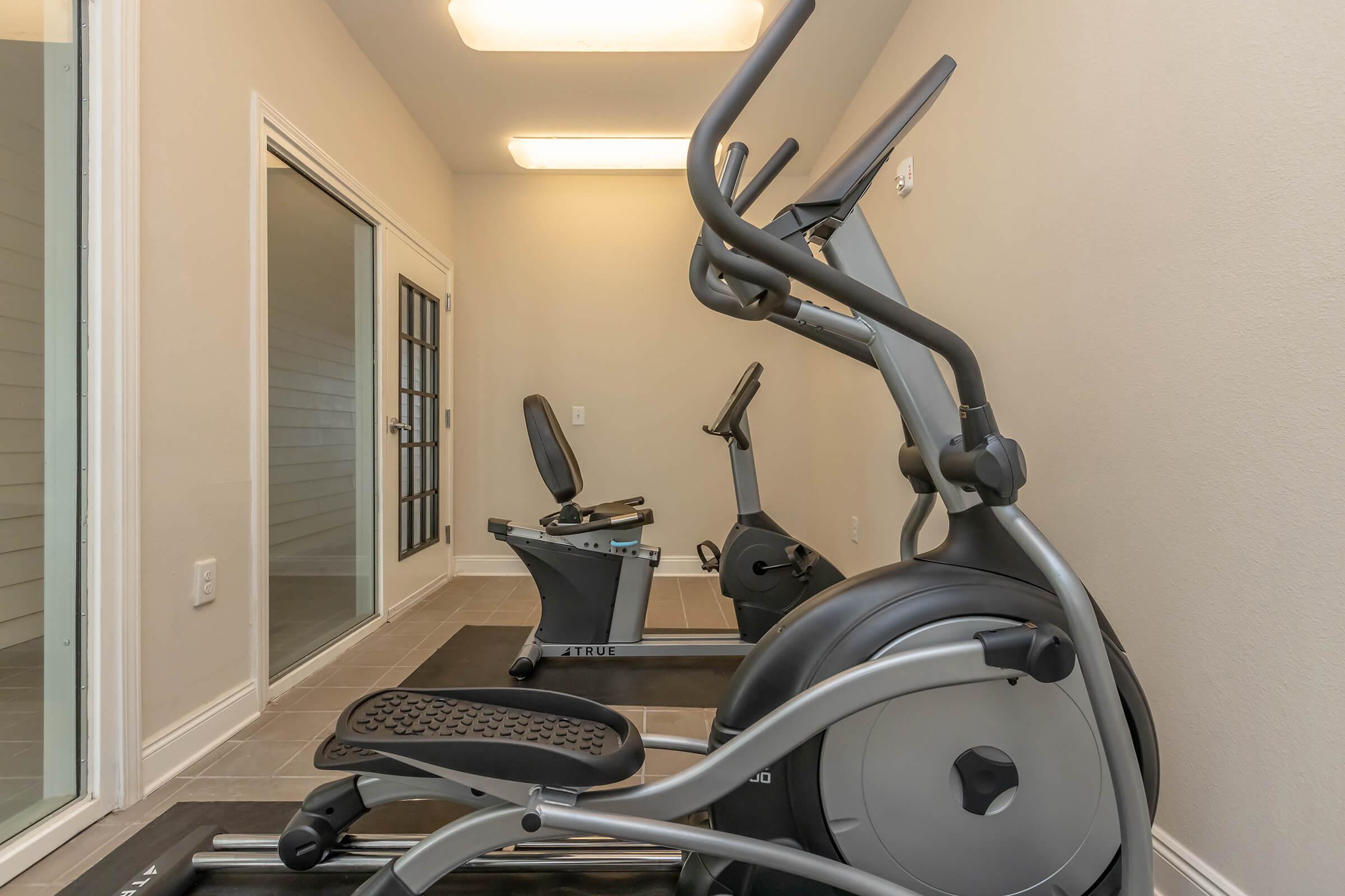 A small exercise room featuring two pieces of gym equipment: an elliptical machine and a stationary bike. The space has neutral-colored walls and tiled flooring, with natural light coming from a nearby door leading outside.