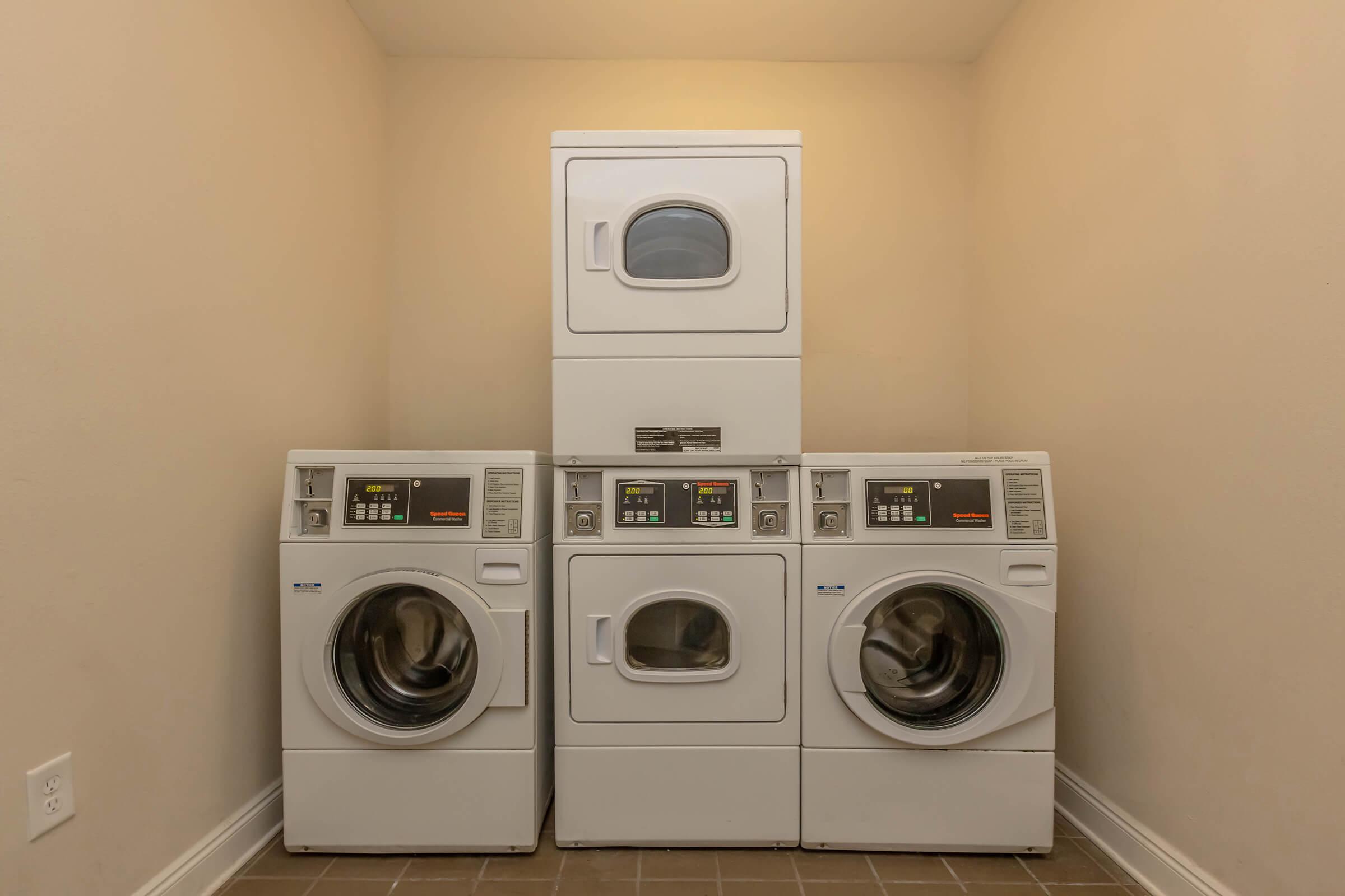 A stacked laundry setup in a neutral-colored room featuring a washing machine on the left, a dryer on the right, and a compact dryer unit on top. The appliances have digital control panels and are situated on a tiled floor. The walls are painted a light color, creating a clean, spacious look.