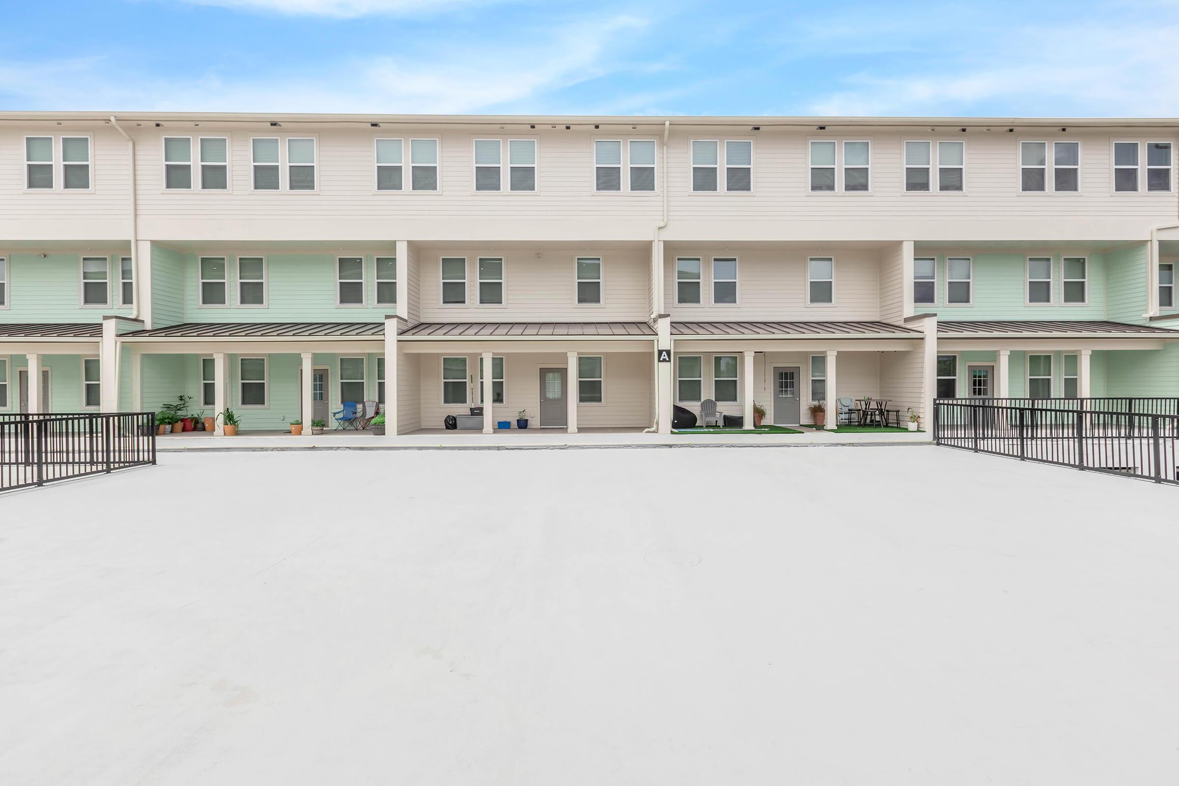 A row of multi-story residential buildings featuring light-colored facades and balconies. The ground area is paved with a smooth surface, surrounded by a black fence. Potted plants and outdoor furniture can be seen on porches. The sky above is blue with a few clouds.