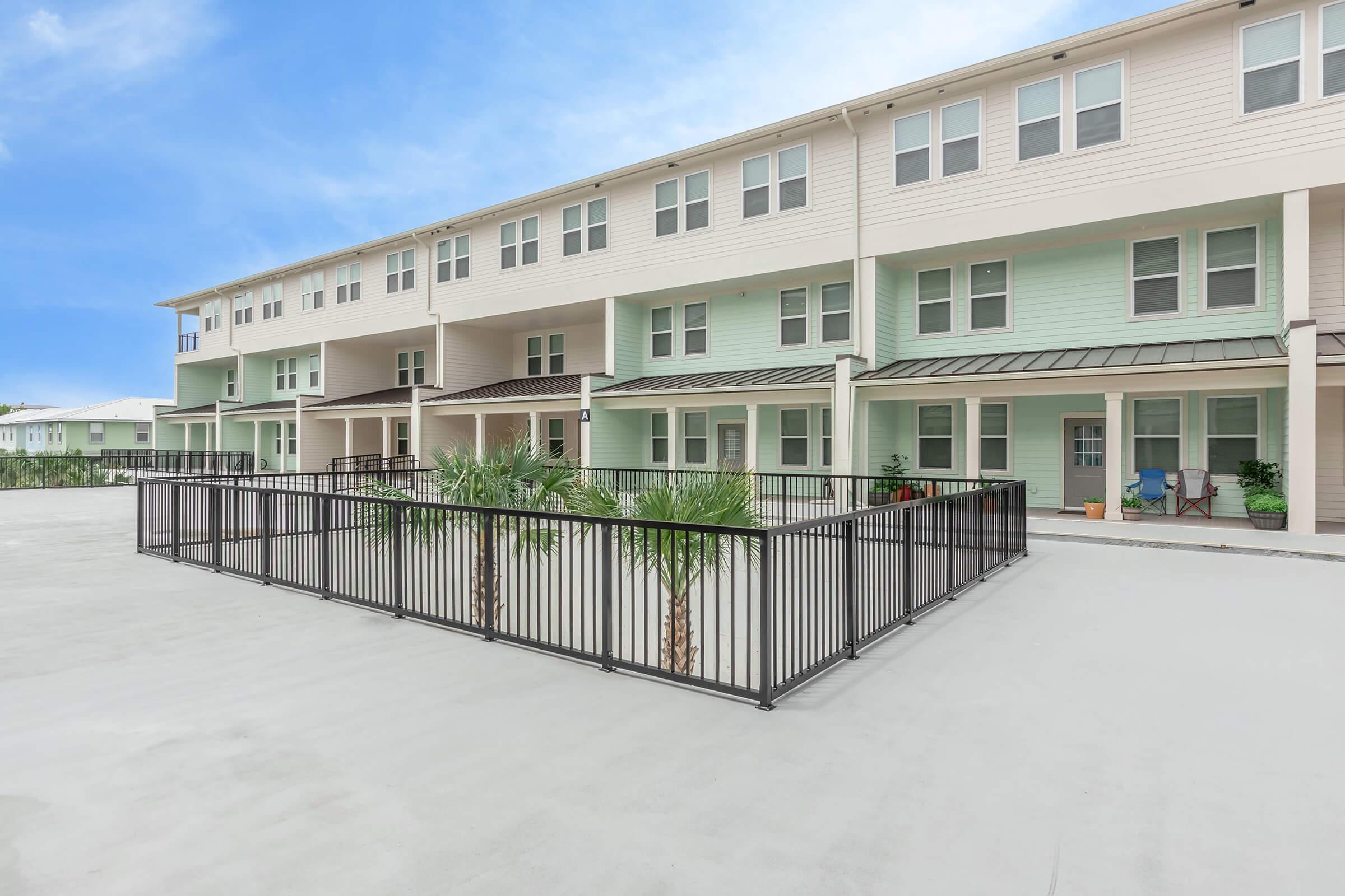 A modern residential building featuring multiple units with a light green and beige exterior. There is a fenced courtyard area with palm trees and clean concrete flooring. The sky is blue with few clouds, suggesting a bright and sunny day.