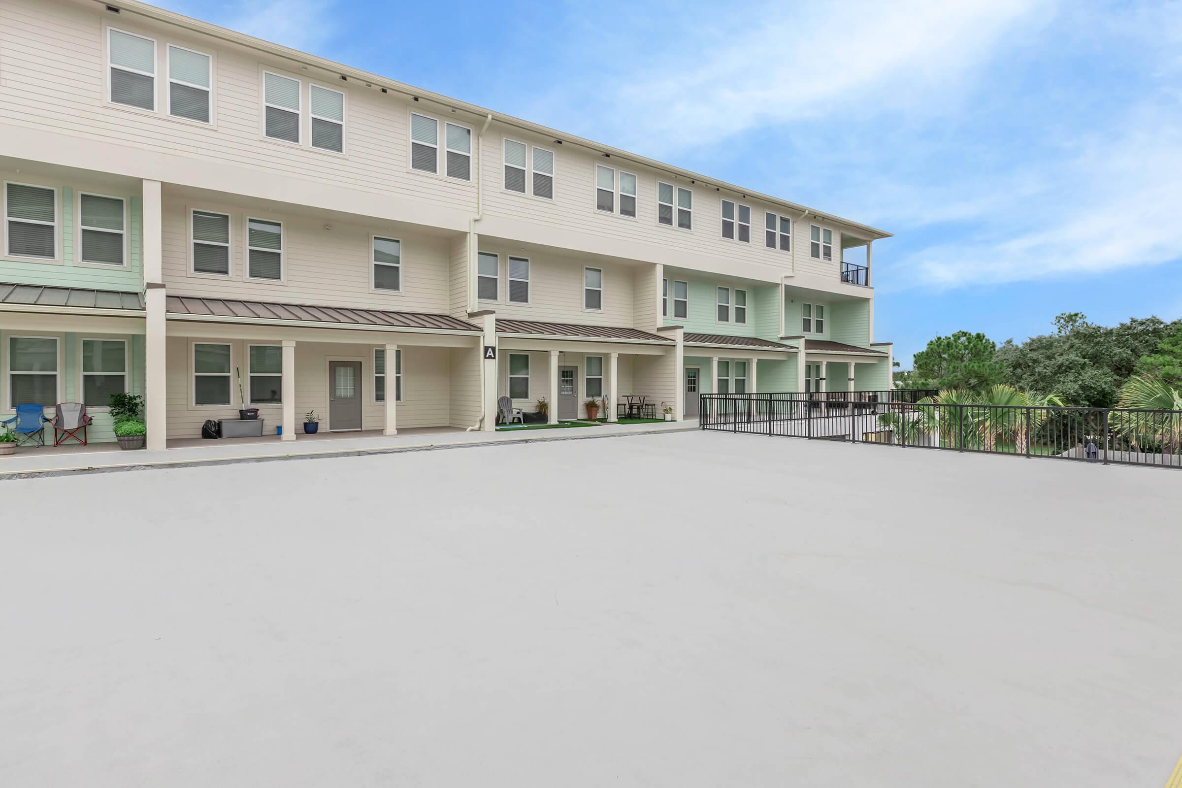 View of a modern apartment complex featuring two-story buildings with multiple windows and a balcony. The exterior includes various neutral-colored siding, a concrete patio area, and landscaping in the background. The sky is clear with soft blue tones, creating a serene atmosphere.