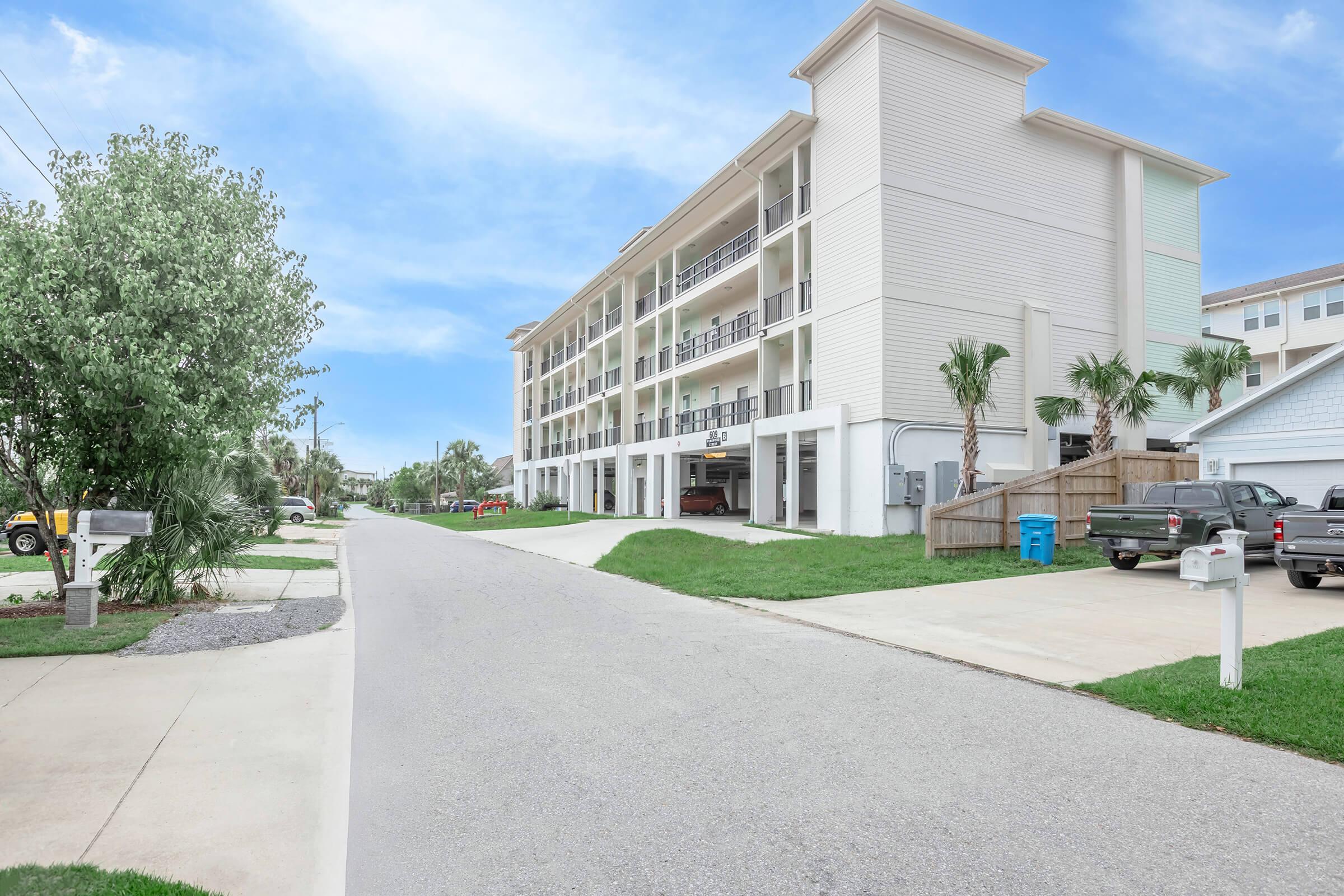 Three-story residential building with a light-colored exterior, featuring balconies on the upper floors. The surrounding area includes green grass and a paved driveway. Palm trees are visible nearby, and two parked cars are on the street. A mailbox stands at the edge of the driveway. Clear blue sky in the background.