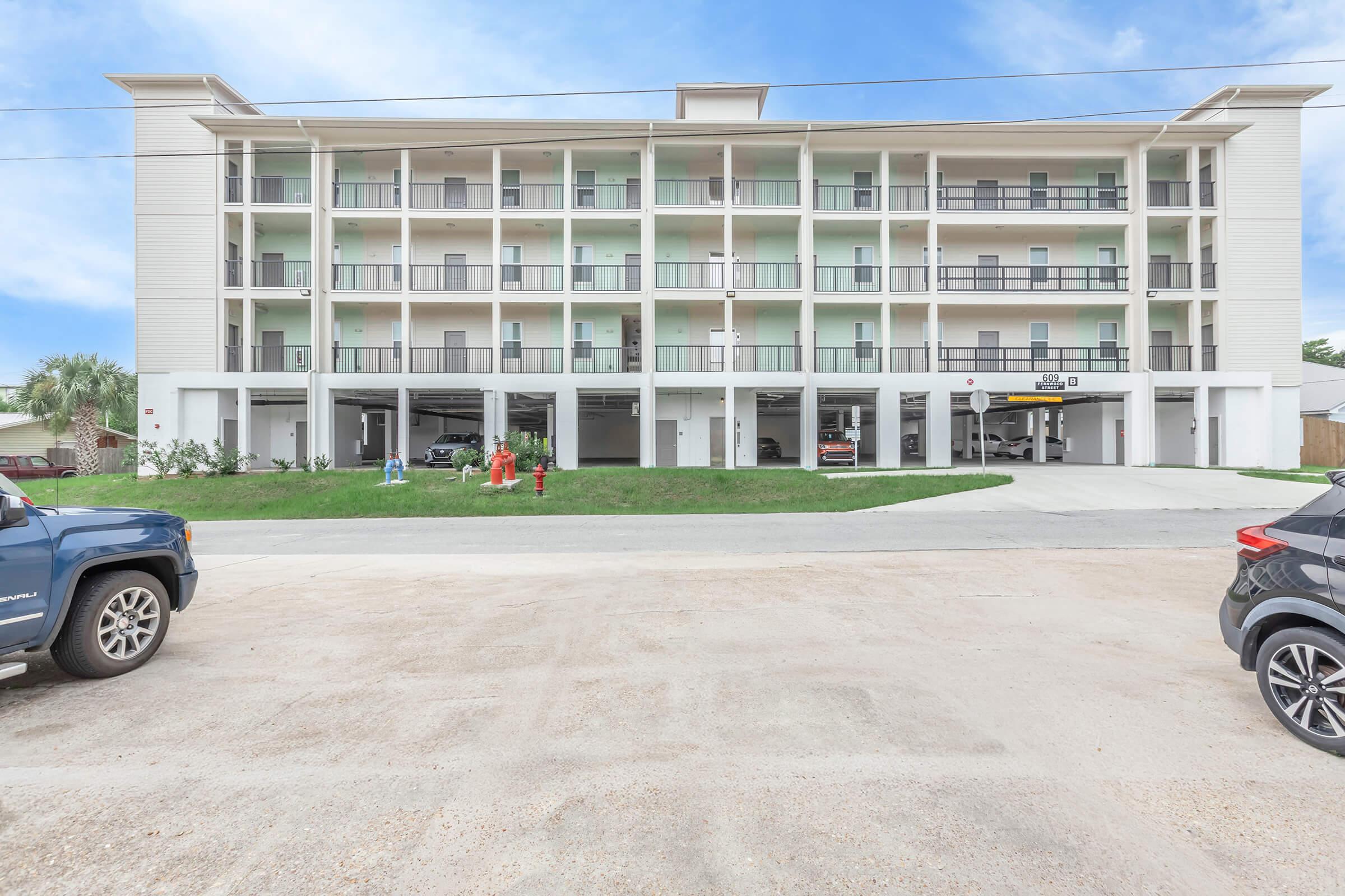 Three-story building featuring multiple balconies and a modern design, with parking spaces in the foreground. A blue vehicle is parked on the left and a black vehicle on the right. In the center, two individuals are engaged in an outdoor activity on the lawn. Clear sky with a few clouds in the background.