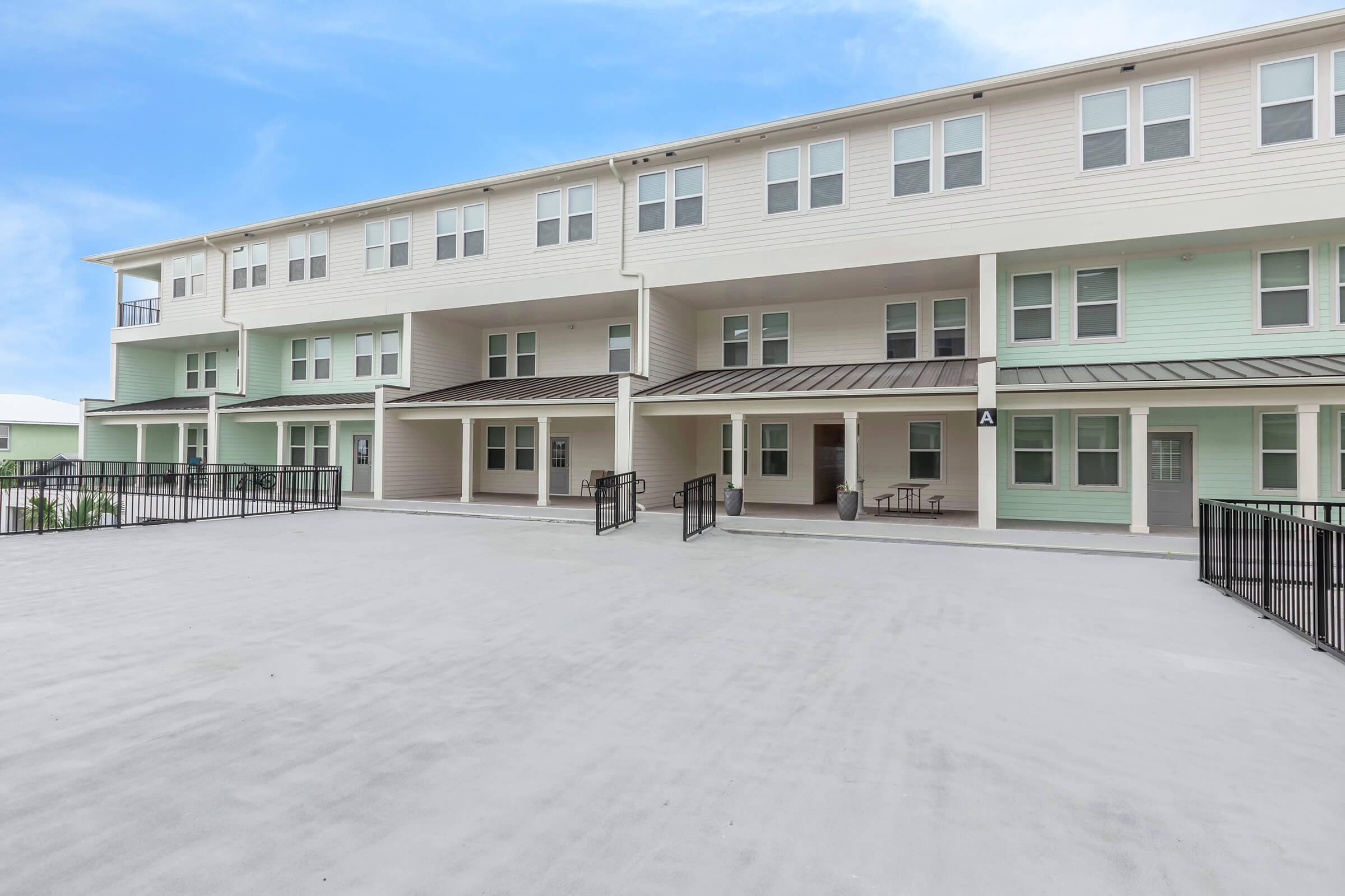 A multi-story residential building with a light-colored exterior. The ground level features a wide concrete patio with black railings and several entrances to individual units. The sky above is clear and blue, enhancing the inviting atmosphere of the space.