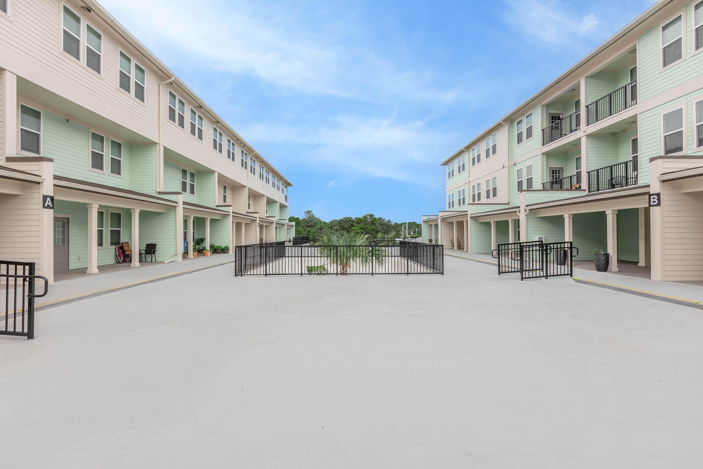 A view of two multi-story residential buildings with light-colored siding on either side of a wide, paved pathway. A black gate is visible in the center, leading to green landscaping beyond. The sky is bright blue with a few clouds.