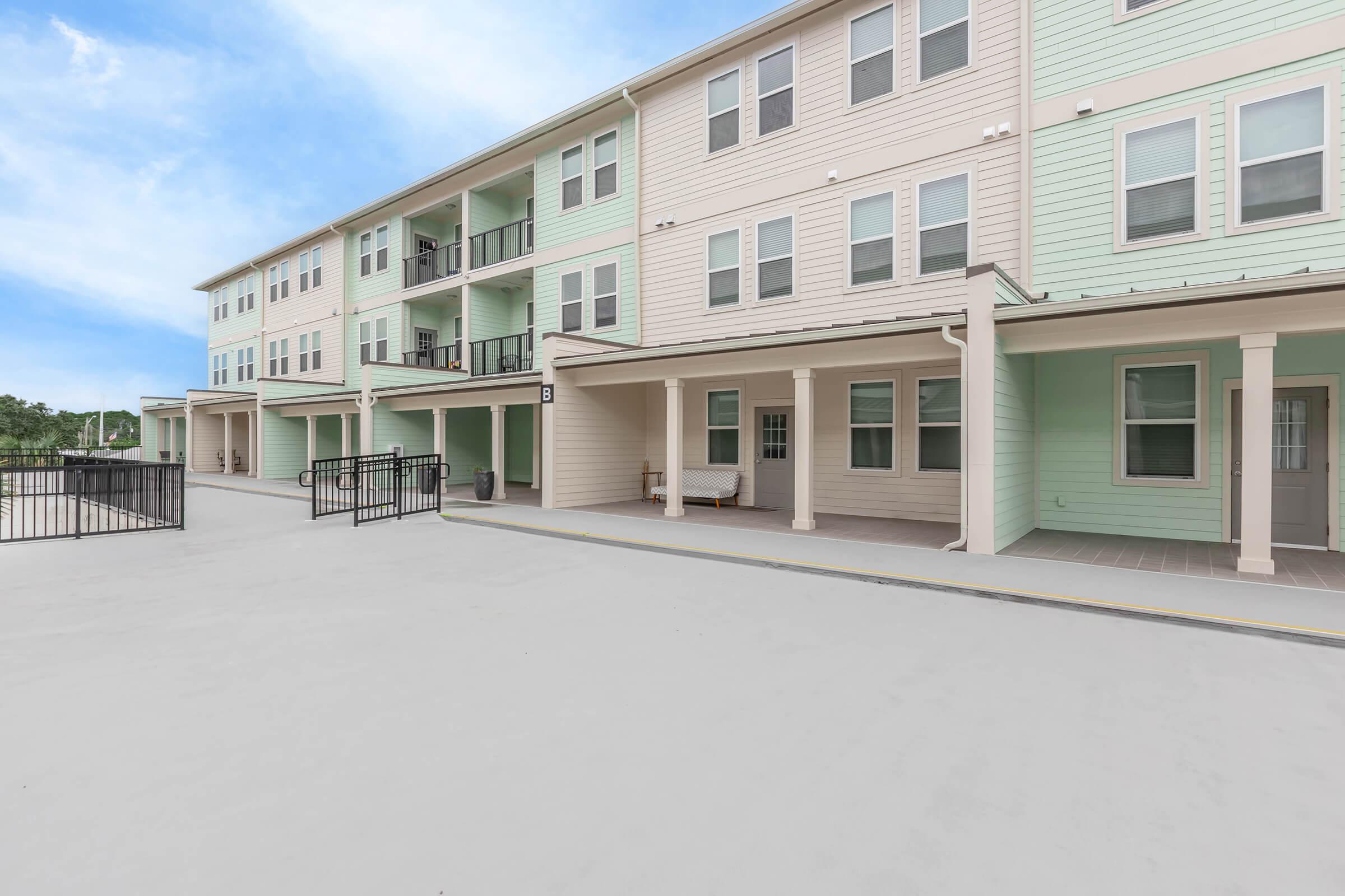 A modern apartment building featuring multiple beige and green units with porches. The area includes a paved surface and a gated entryway, with some outdoor seating visible. The sky is clear and blue in the background, creating a pleasant and welcoming atmosphere.