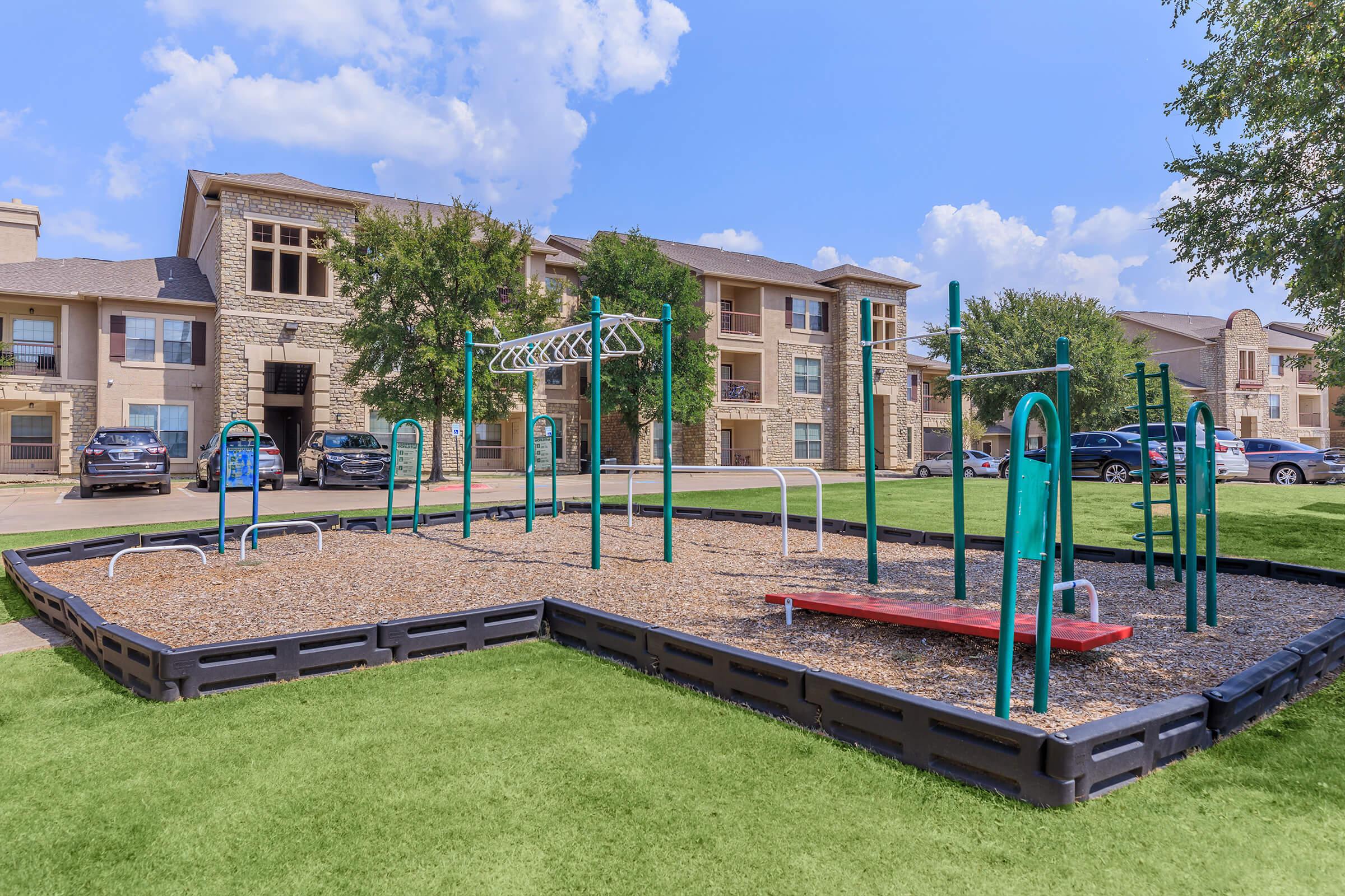 A playground with various equipment, including pull-up bars and slides, surrounded by grass. In the background, there are residential buildings with several parked cars. The sky is blue with a few clouds, creating a bright and inviting atmosphere.
