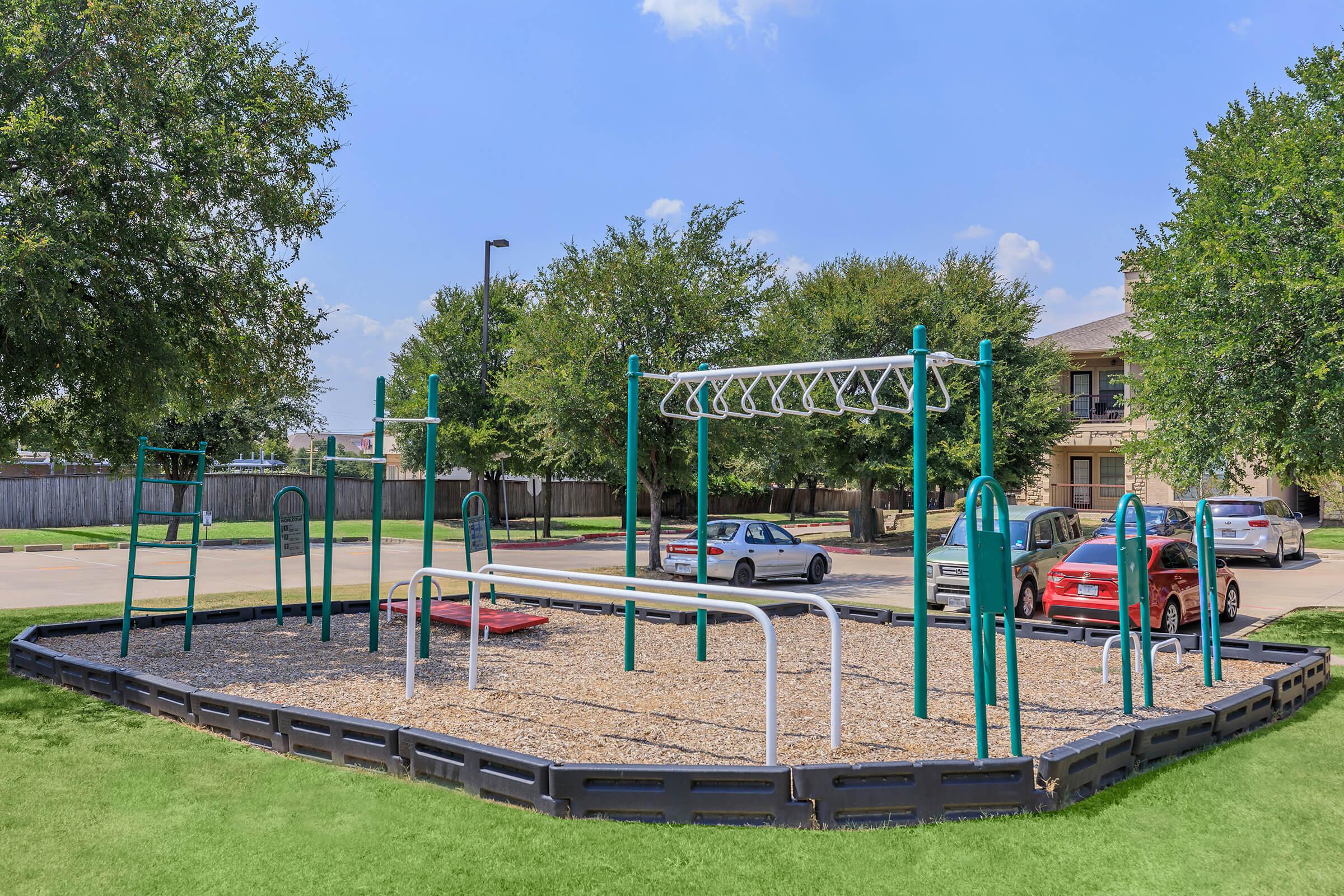 A playground featuring swings, climbing structures, and monkey bars, surrounded by a grassy area. Nearby, cars are parked along a driveway, and trees provide shade in the background. The scene is bright and sunny, indicating a pleasant day for outdoor activities.