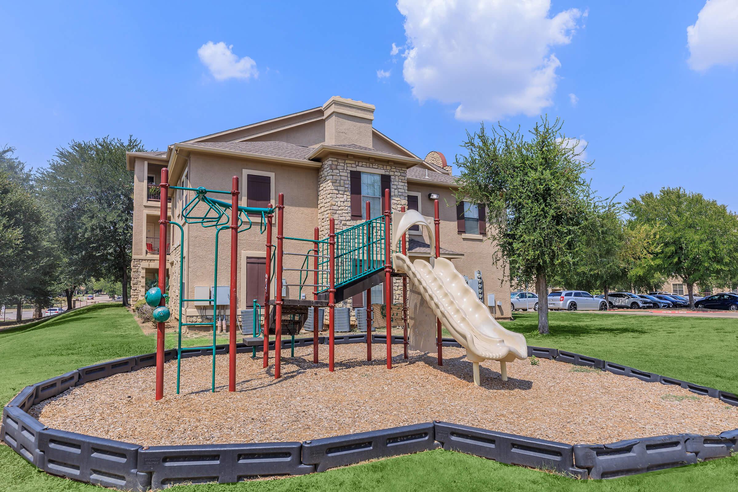 A playground featuring a slide, climbing structures, and swings, set on a grassy area with wood chip ground cover. In the background, a multi-story residential building is visible, surrounded by trees and parked cars. The sky is blue with a few clouds.