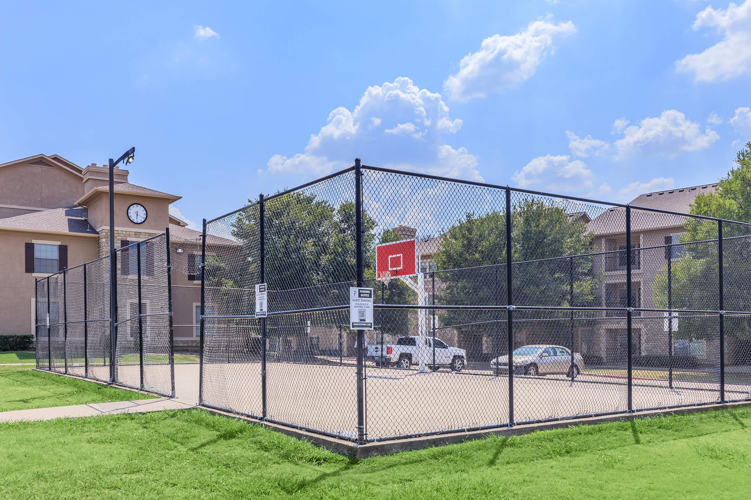 Basketball court enclosed by a chain-link fence, located near residential buildings. The court features a basketball hoop at the far end and is surrounded by green grass and trees under a partly cloudy blue sky. A few parked cars are visible nearby.