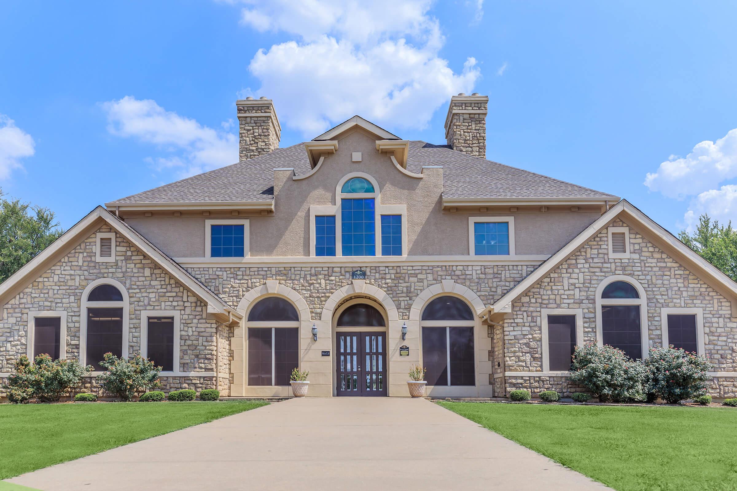 A large, elegant house featuring a stone exterior, multiple windows, and a grand entrance. Surrounded by well-maintained landscaping, the home has a symmetrical design with two prominent chimneys and a spacious front porch, under a bright blue sky with fluffy clouds.