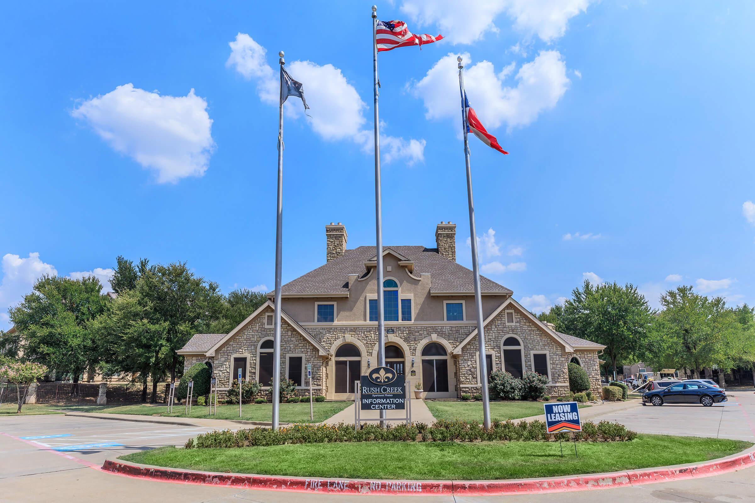A large stone building with a peaked roof, featuring multiple windows and a welcoming entrance. Three flagpoles display the American flag, a state flag, and another flag. A sign in front reads "Now Leasing," and the surrounding area includes green trees and a clear blue sky with fluffy clouds.