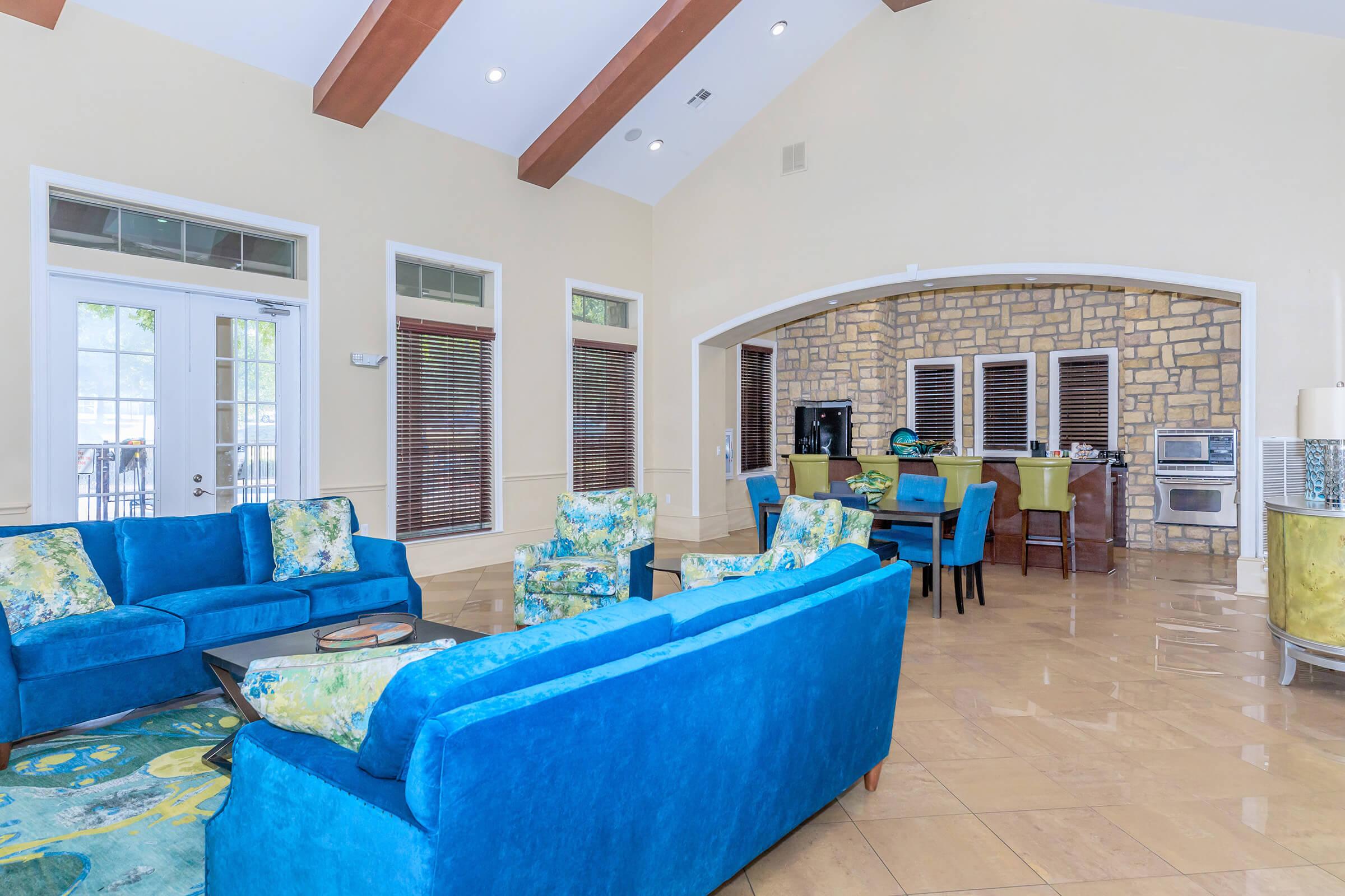 Bright and airy living room featuring blue sofas and a floral patterned chair set. Stone accents on one wall and arched entry to the kitchen area. Large windows allow natural light, and pale yellow walls enhance the spacious feel. Modern decor with an inviting atmosphere.