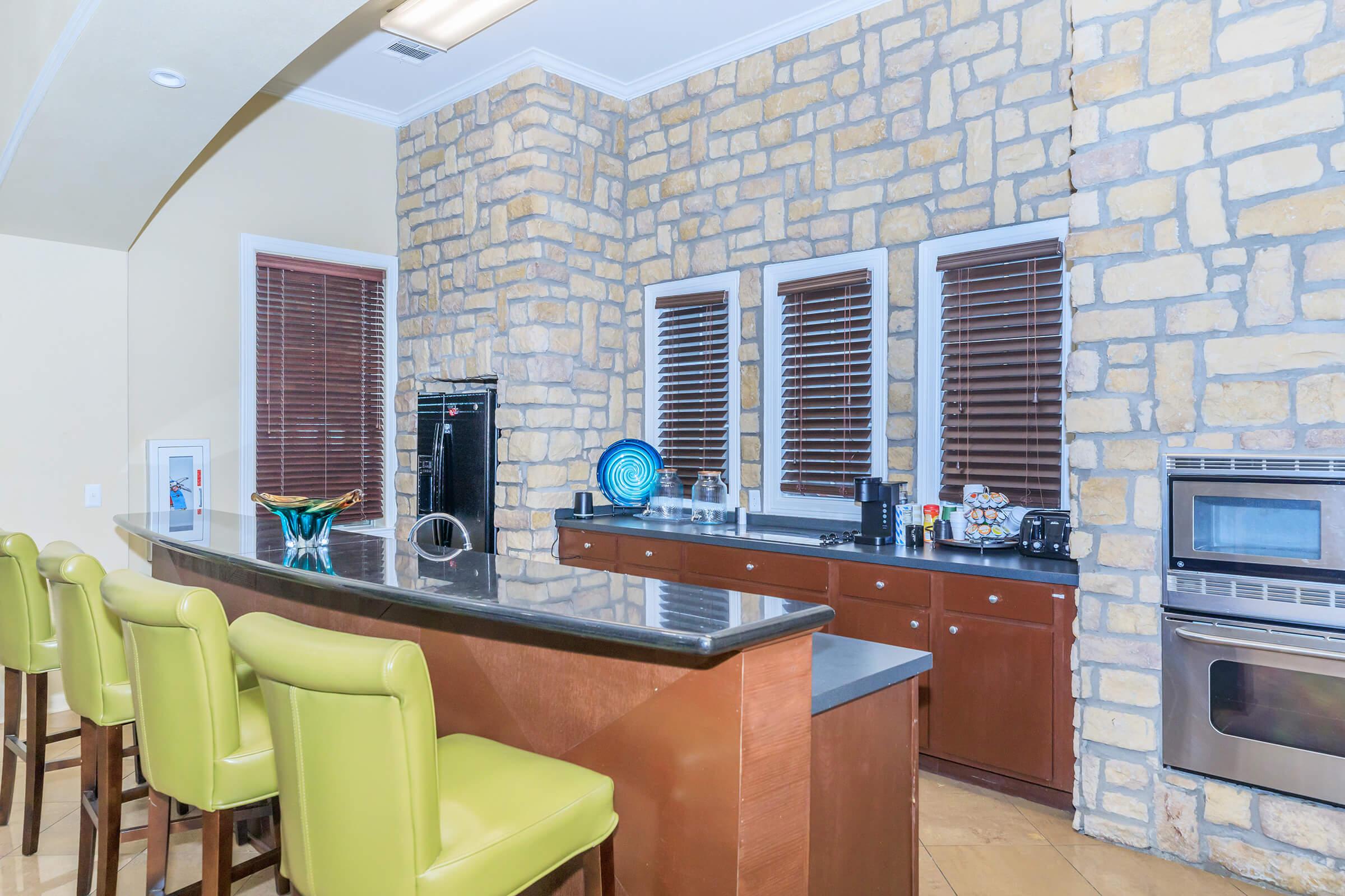 Modern kitchen featuring a stone wall, dark wood cabinets, black countertop, and stainless steel appliances. Bar seating with green chairs is arranged at the counter. Large windows with wooden blinds provide natural light. Kitchen appliances and decorative items are neatly displayed on the counter.