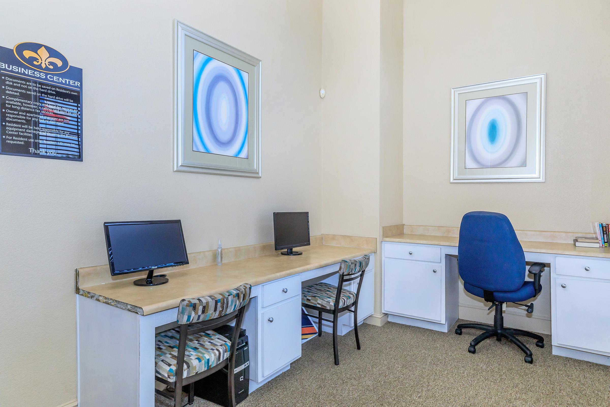A business center with two desks featuring monitors, a blue office chair, and light-colored walls. There are two abstract art pieces in decorative frames on the wall, and informational signage in the corner. The space is bright and organized, designed for productivity.