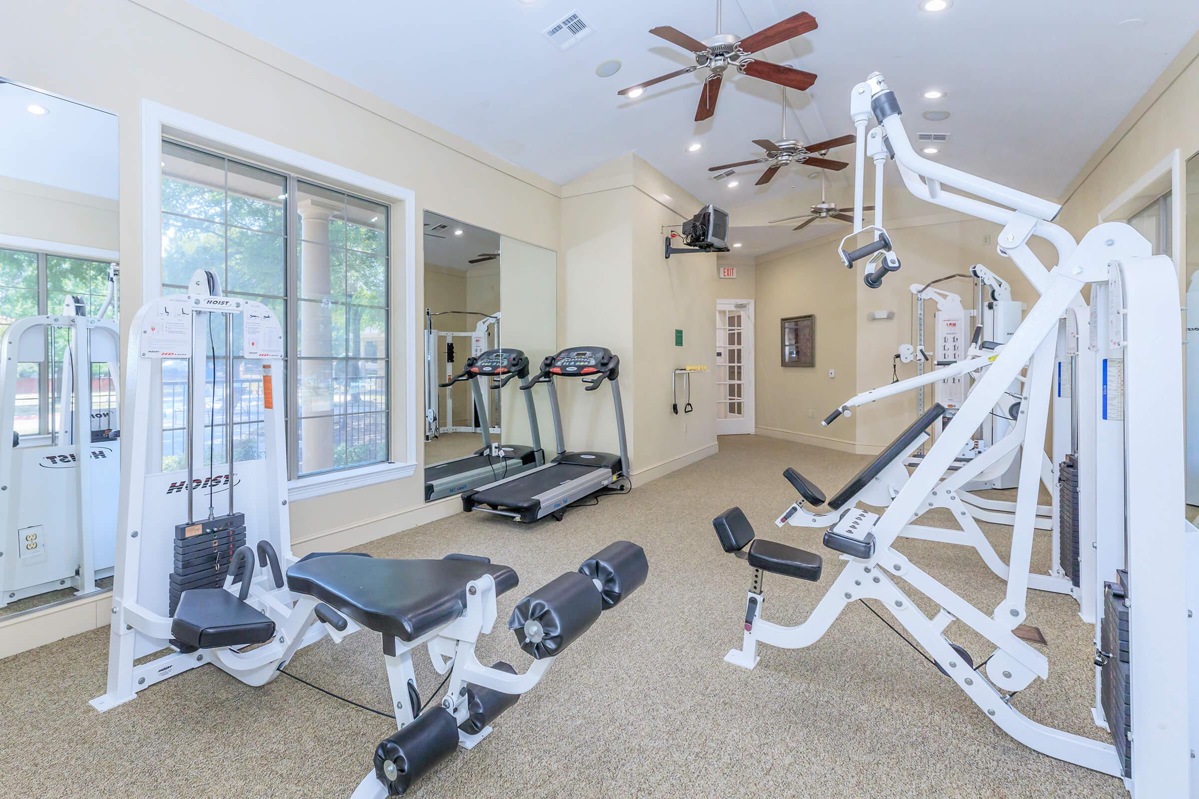 A spacious gym area featuring various exercise equipment, including treadmills, weight machines, and an exercise bench. The room has large windows allowing natural light, ceiling fans for ventilation, and a neutral-colored carpet, creating a bright and inviting fitness space.