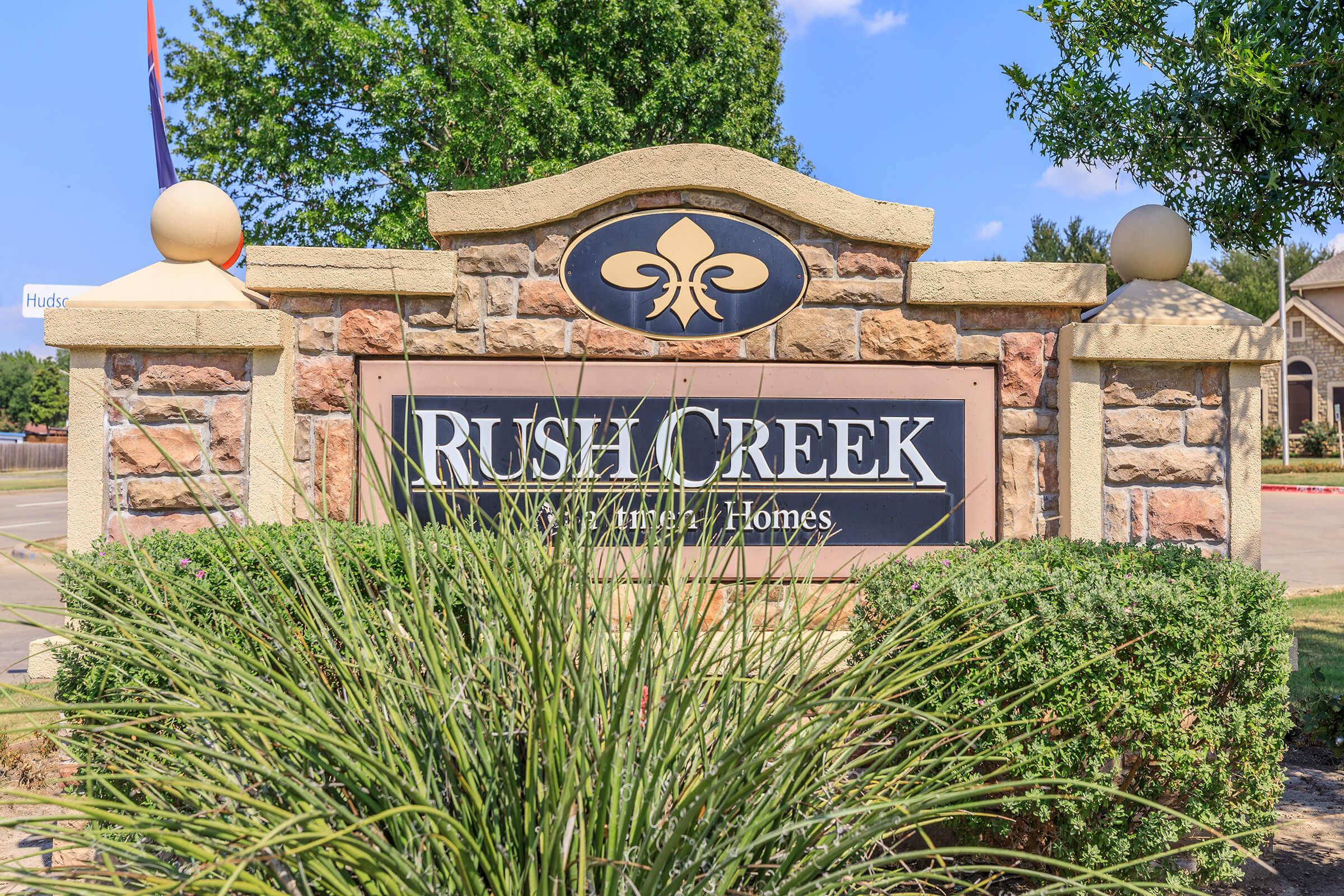 Sign for Rush Creek New Urban Homes, featuring a decorative stone structure with a dark sign that displays the name "Rush Creek." Surrounding greenery includes grass and shrubs, with a blue sky and some trees in the background.