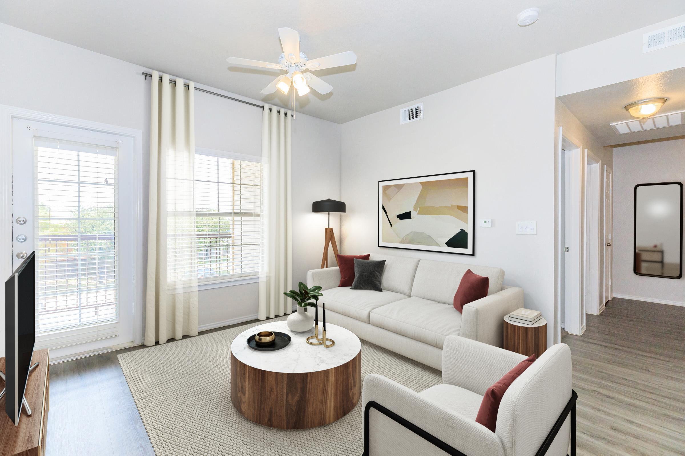 Modern living room featuring a light-colored sofa with red accents, a round coffee table, and a ceiling fan. There's a window with blinds and sheer curtains letting in natural light. A flat-screen TV is placed on a wooden stand, and a minimalist artwork is displayed on the wall.