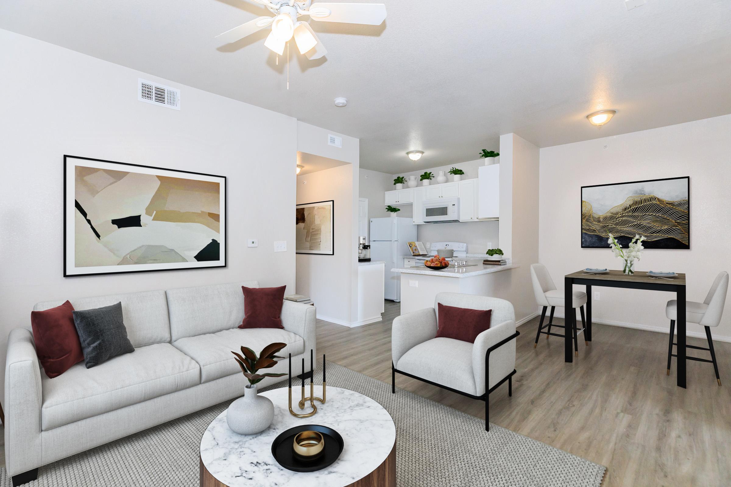 A modern living room with a light-colored sofa adorned with red and black pillows. There's a round coffee table with a decorative plant centerpiece. In the background, a dining area features a small table with white chairs. The kitchen is visible with white cabinets and appliances, and the walls are painted in soft neutral tones.
