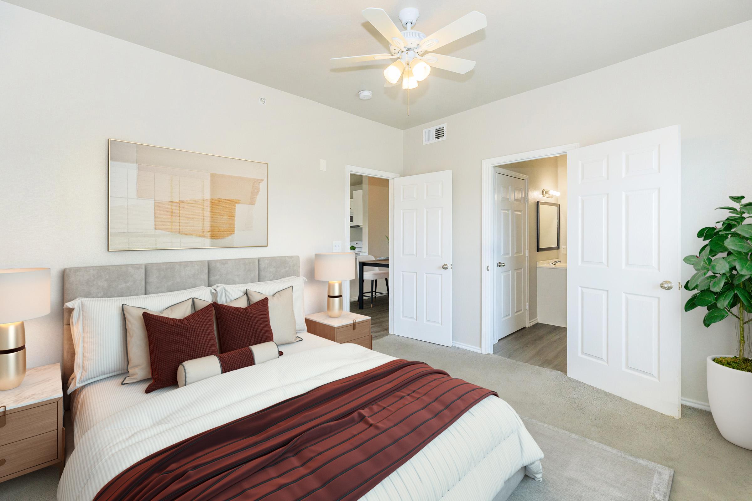 A cozy bedroom featuring a comfortable bed with a grey headboard and decorative pillows, two bedside lamps, and light-colored walls. The room includes a ceiling fan and a potted plant. Two doors lead to separate areas, one presumably to a bathroom. The space is well-lit and styled for a modern aesthetic.