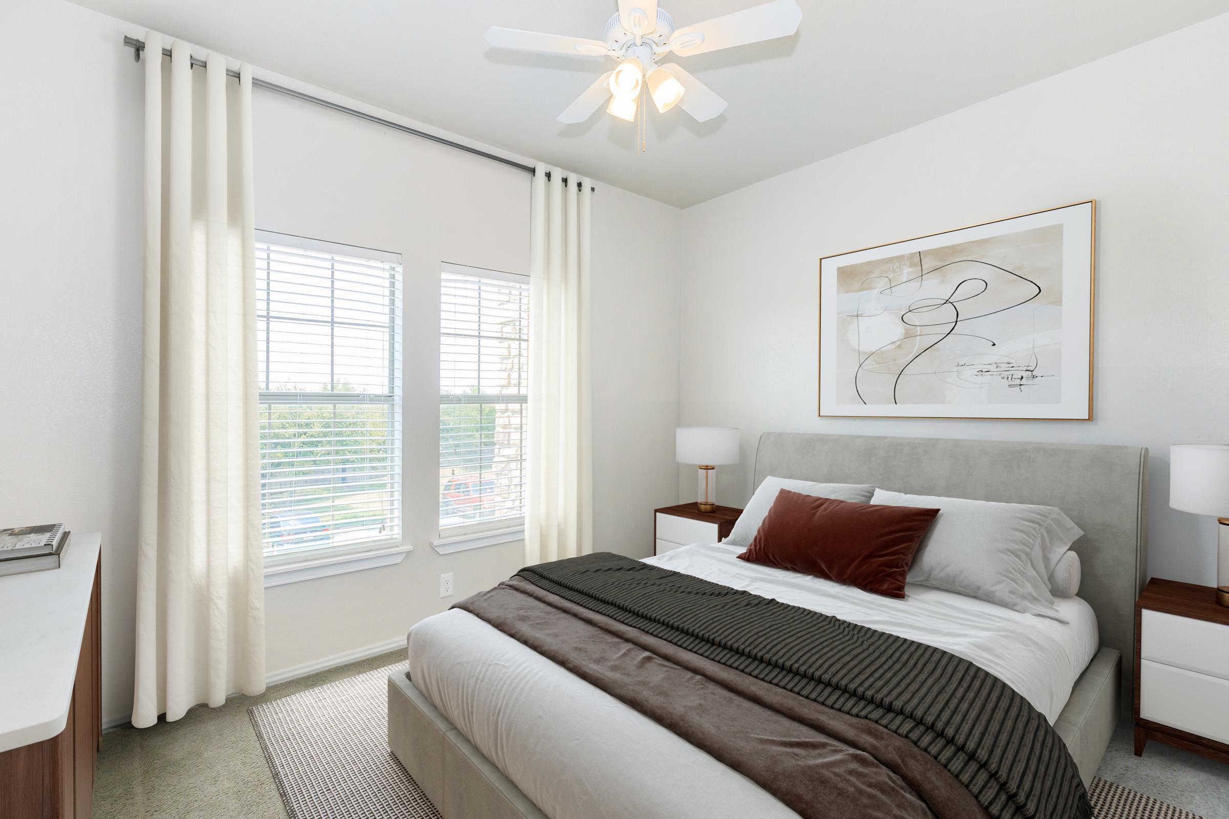 A cozy bedroom featuring a neatly made bed with decorative pillows, a bedside table with lamps on either side, a light-colored rug, and large windows with sheer curtains allowing natural light to fill the space. The walls are painted in a soft color, and there’s a modern artwork hanging above the bed.