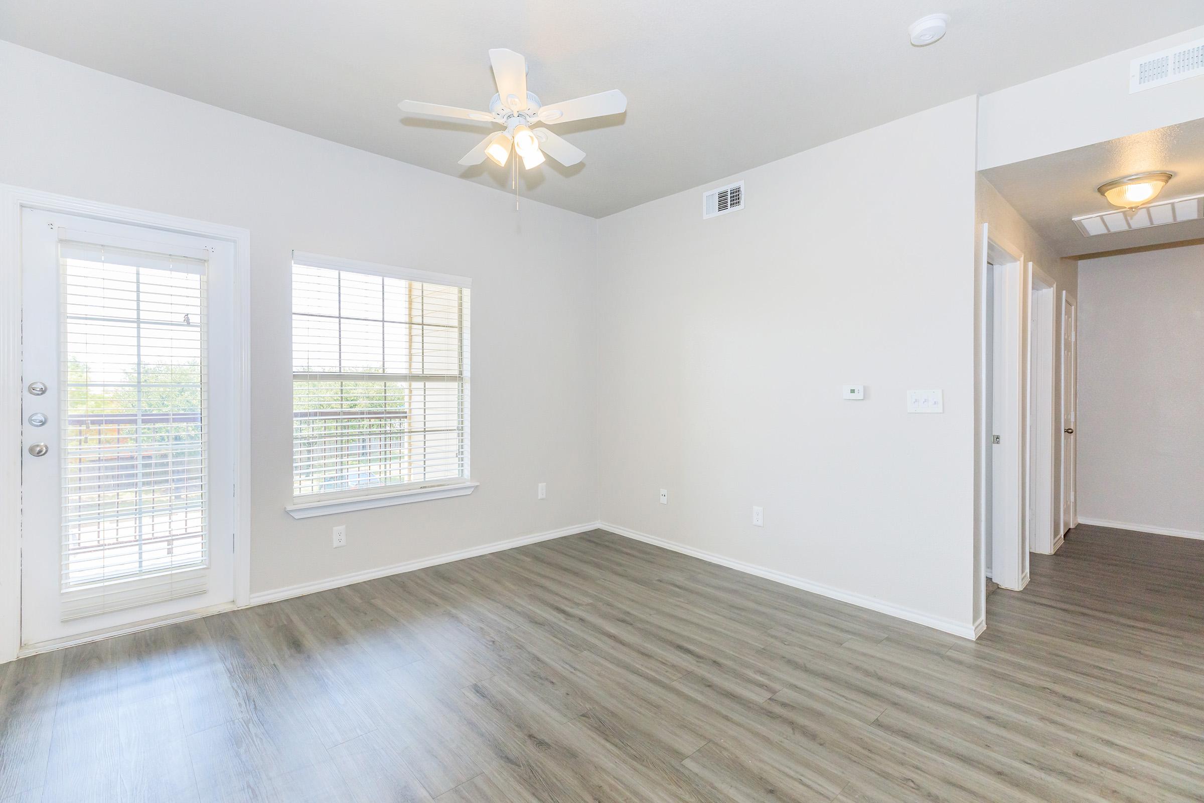 Interior of a bright, minimalistic room featuring light gray walls, a ceiling fan, and modern wood-like flooring. One wall has a sliding glass door leading to a balcony, and there are two windows with blinds allowing natural light. A hallway is visible to the right, leading to other areas.