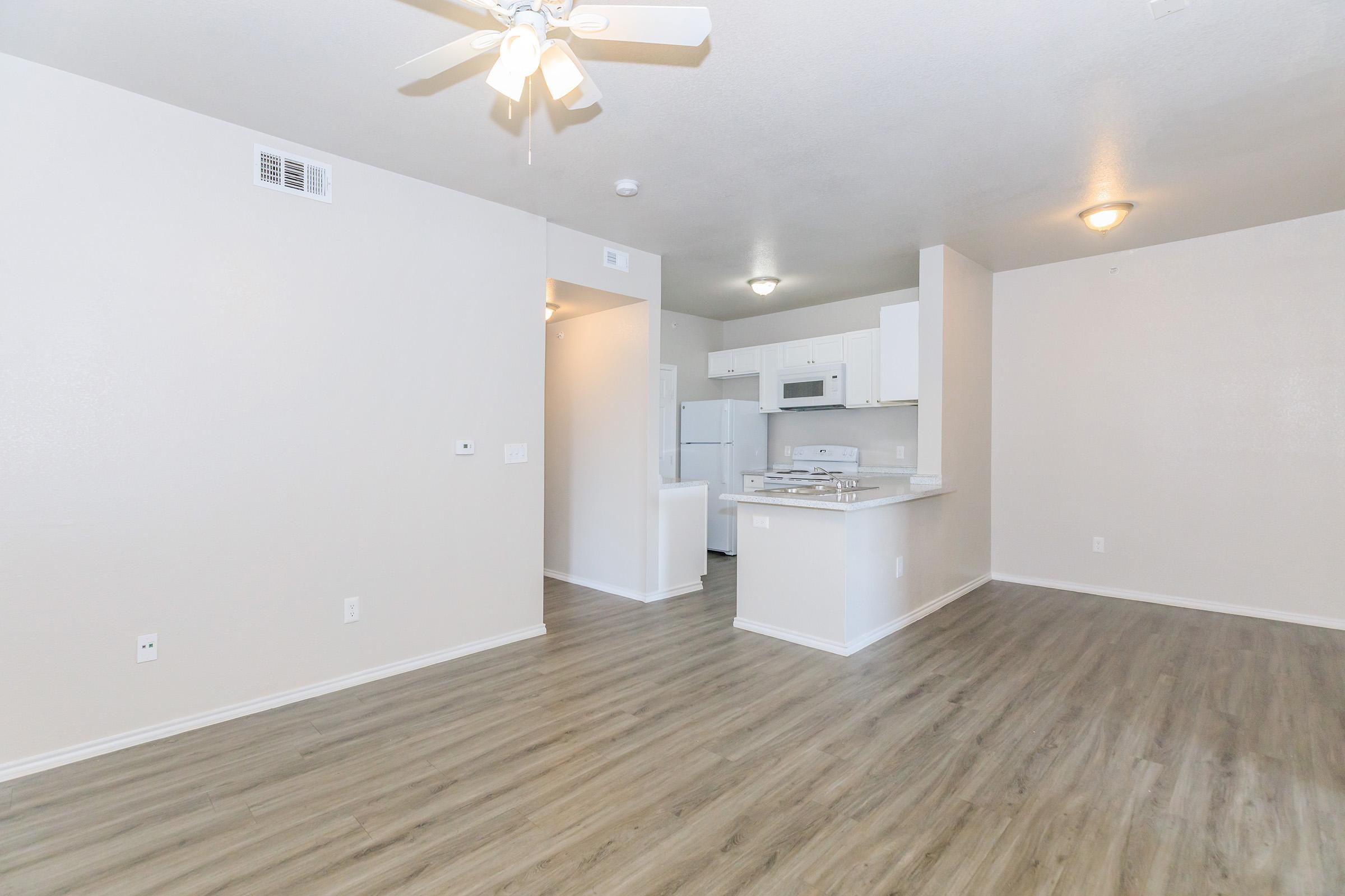 Bright and spacious apartment interior featuring light-colored walls, laminate flooring, and a ceiling fan. The open layout includes a view of the kitchen with white cabinetry and appliances, creating an inviting living space.